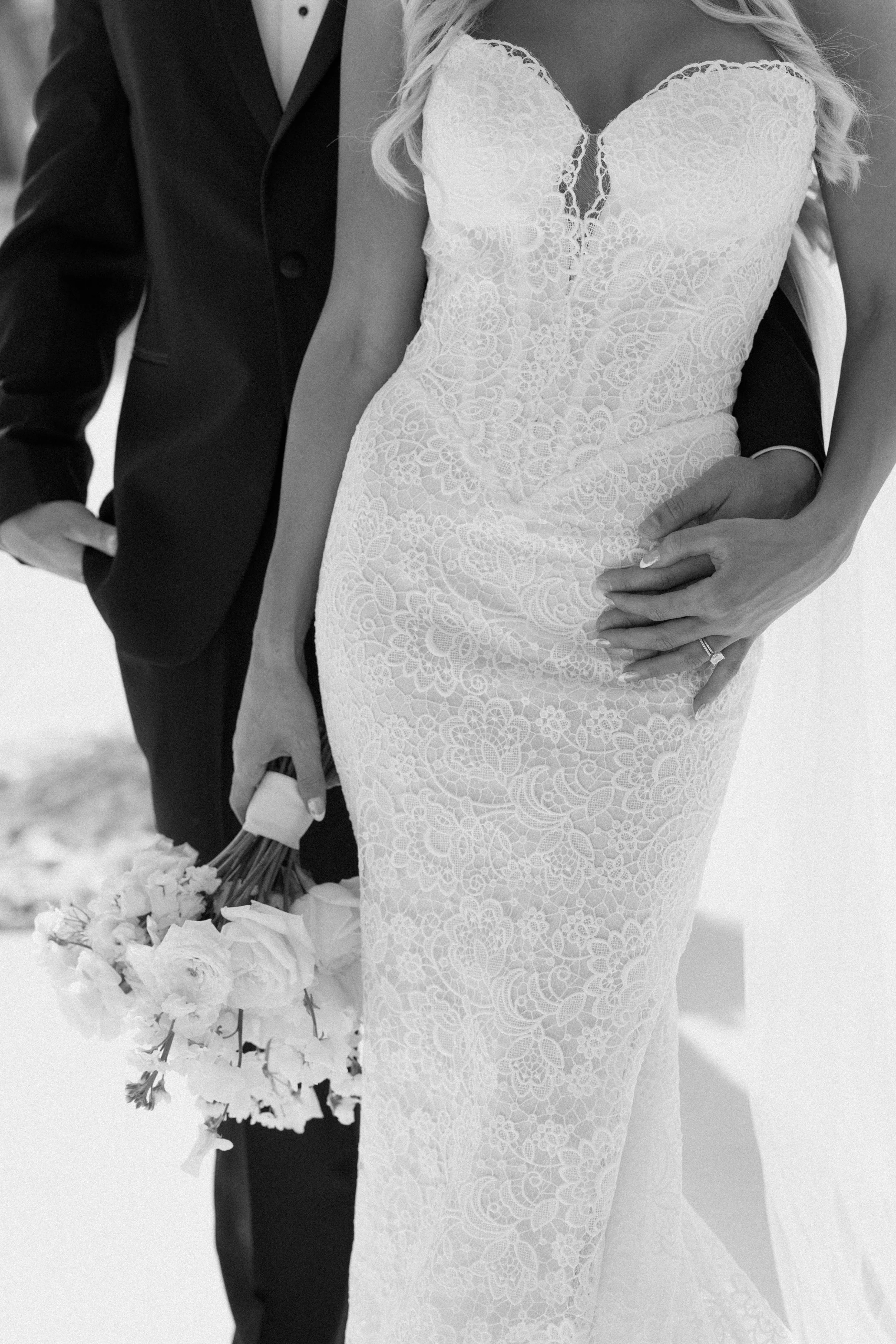 Black and white photo of a bride and groom, with the bride wearing a lace wedding dress and holding a bouquet of flowers, and the groom dressed in a black tuxedo.