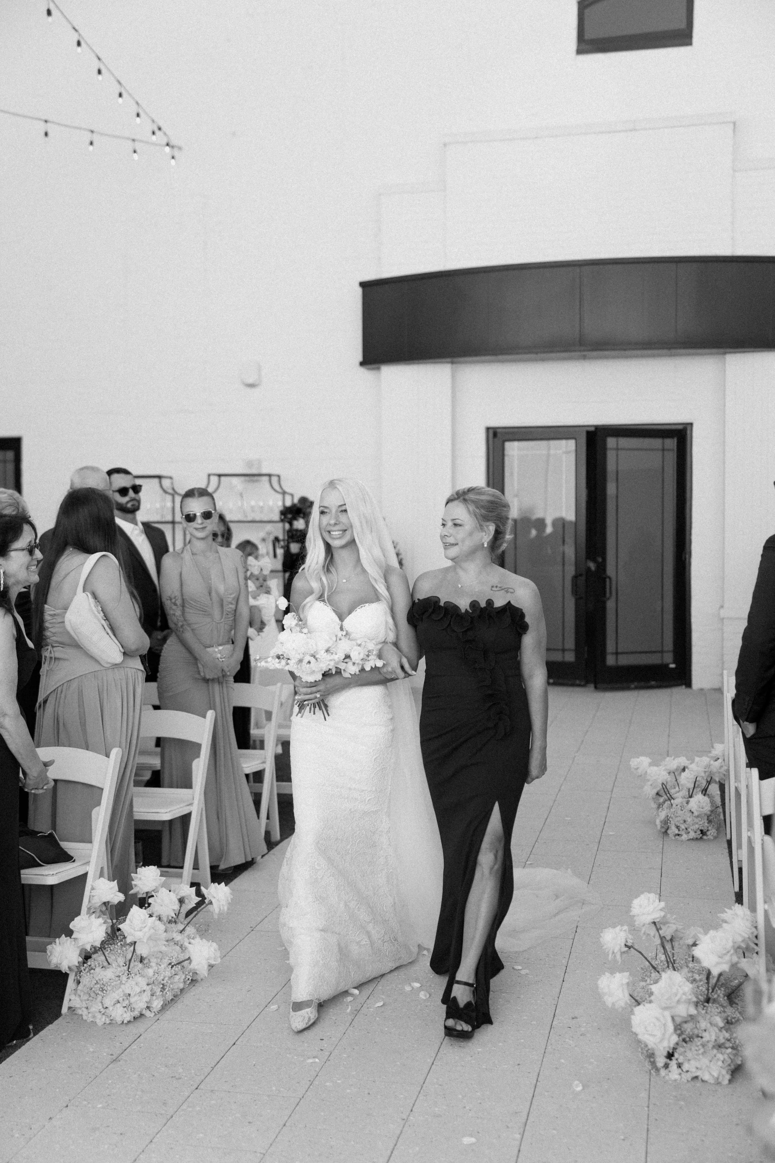 A bride in a white lace wedding dress holding a bouquet, walking down the aisle with a woman in a black dress at her side, at an outdoor wedding ceremony. Guests are seated on either side, with some standing and wearing formal attire.