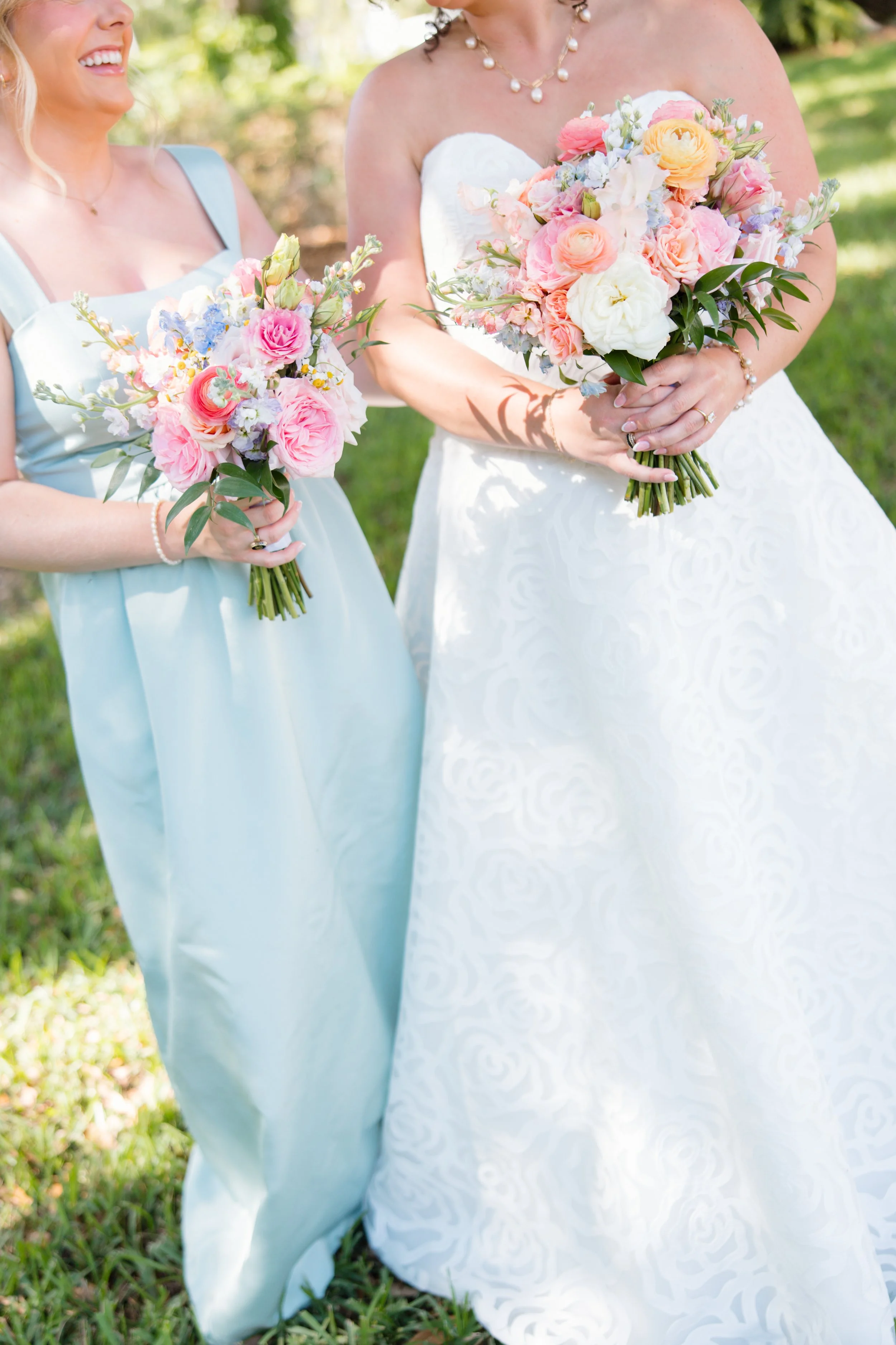 Close-up of two women holding bouquets of pink, white, and pastel flowers, wearing wedding attire outdoors.