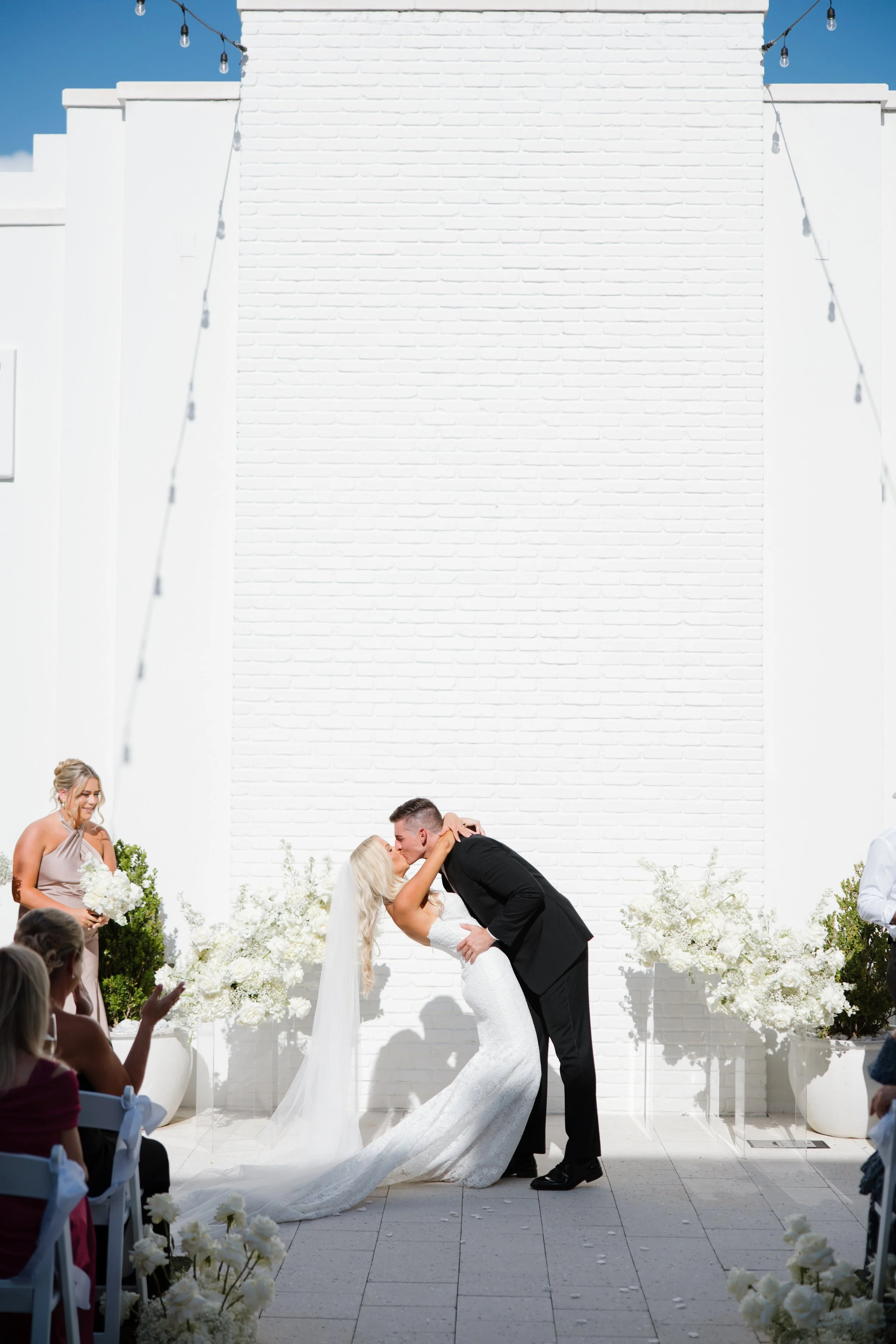 A bride and groom kiss during their outdoor wedding ceremony, with guests seated in white chairs, a large white brick wall in the background, and floral arrangements along the aisle.