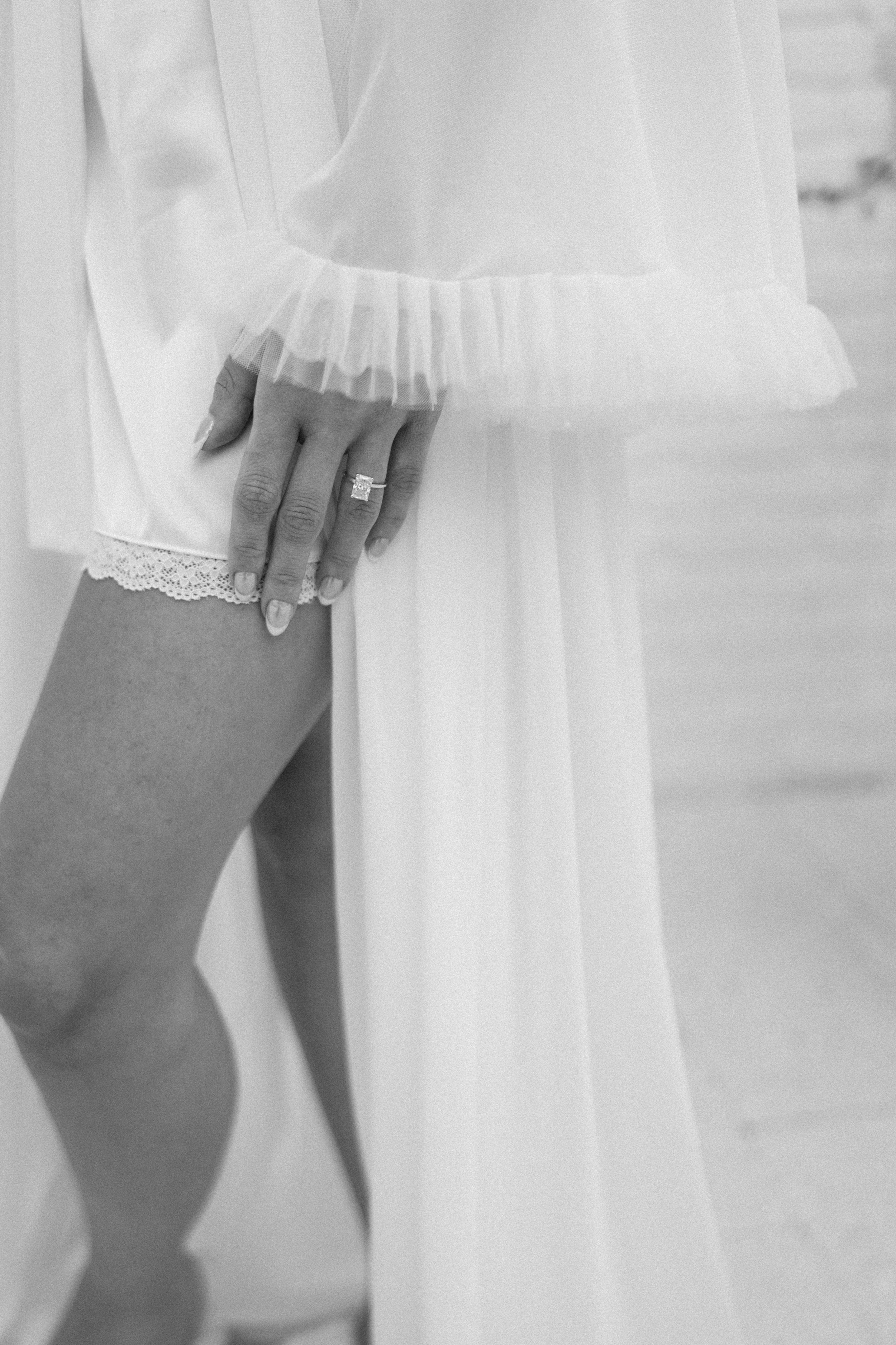 A black-and-white photo of a person wearing a sheer, long-sleeved tulle top with ruffle cuffs, a lace-trimmed slip or dress underneath, and a diamond ring on their finger. The person is resting their hand on their thigh.
