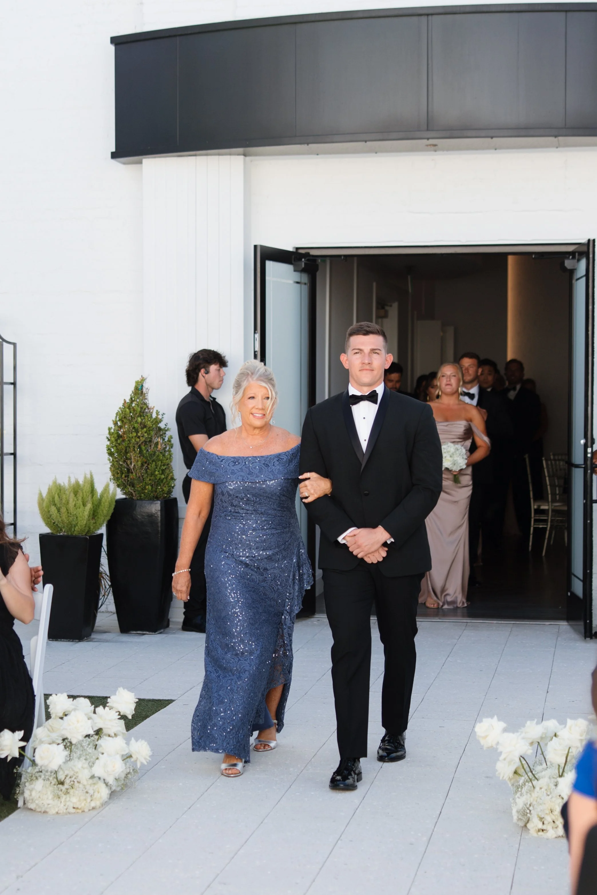 A bride and groom walking down an aisle at their wedding ceremony. The bride is wearing a blue lace gown, and the groom is in a black tuxedo. They are outside a building with guests and bridesmaids in the background.