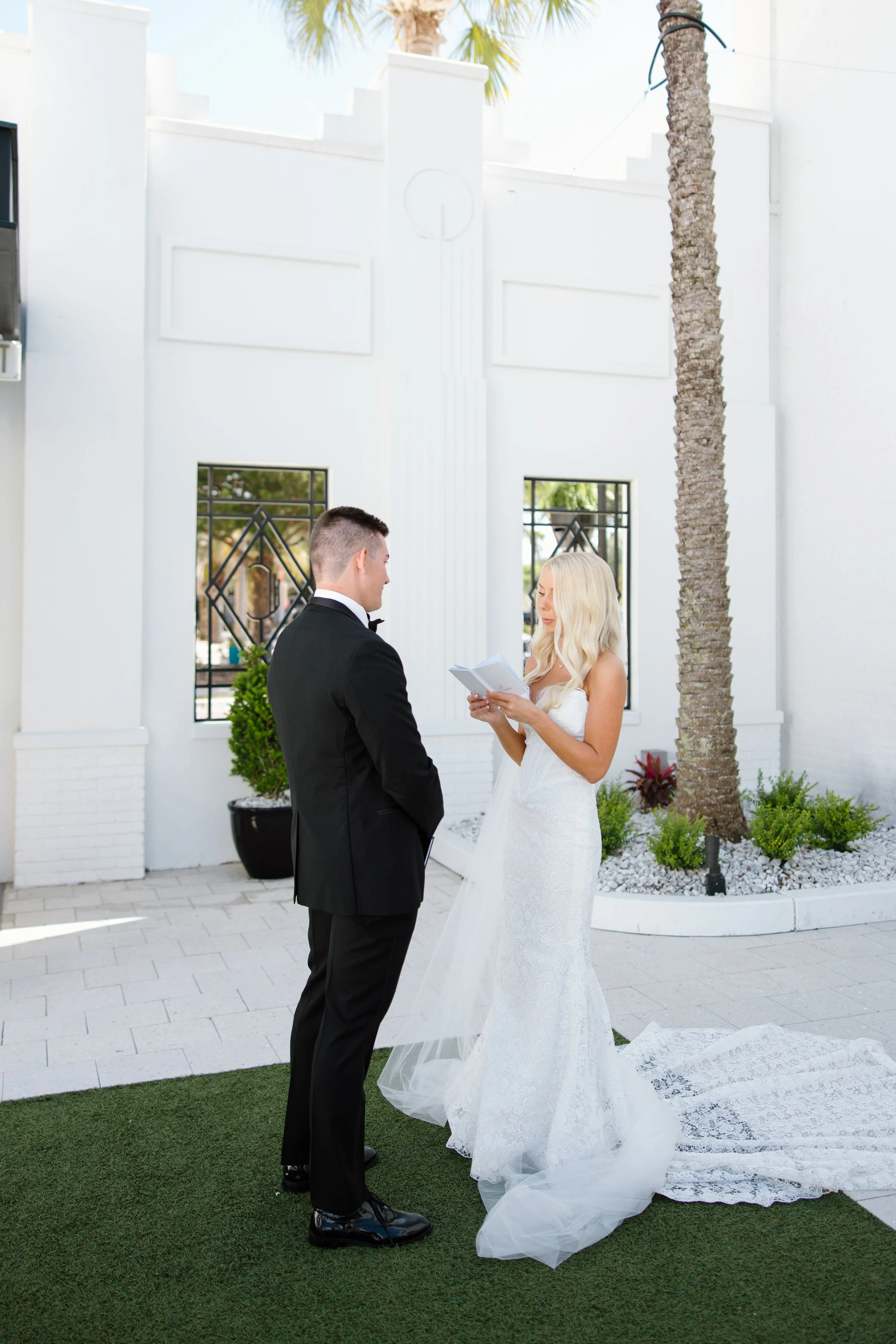 A bride and groom exchanging vows outdoors in front of a white modern building with large windows and palm trees.