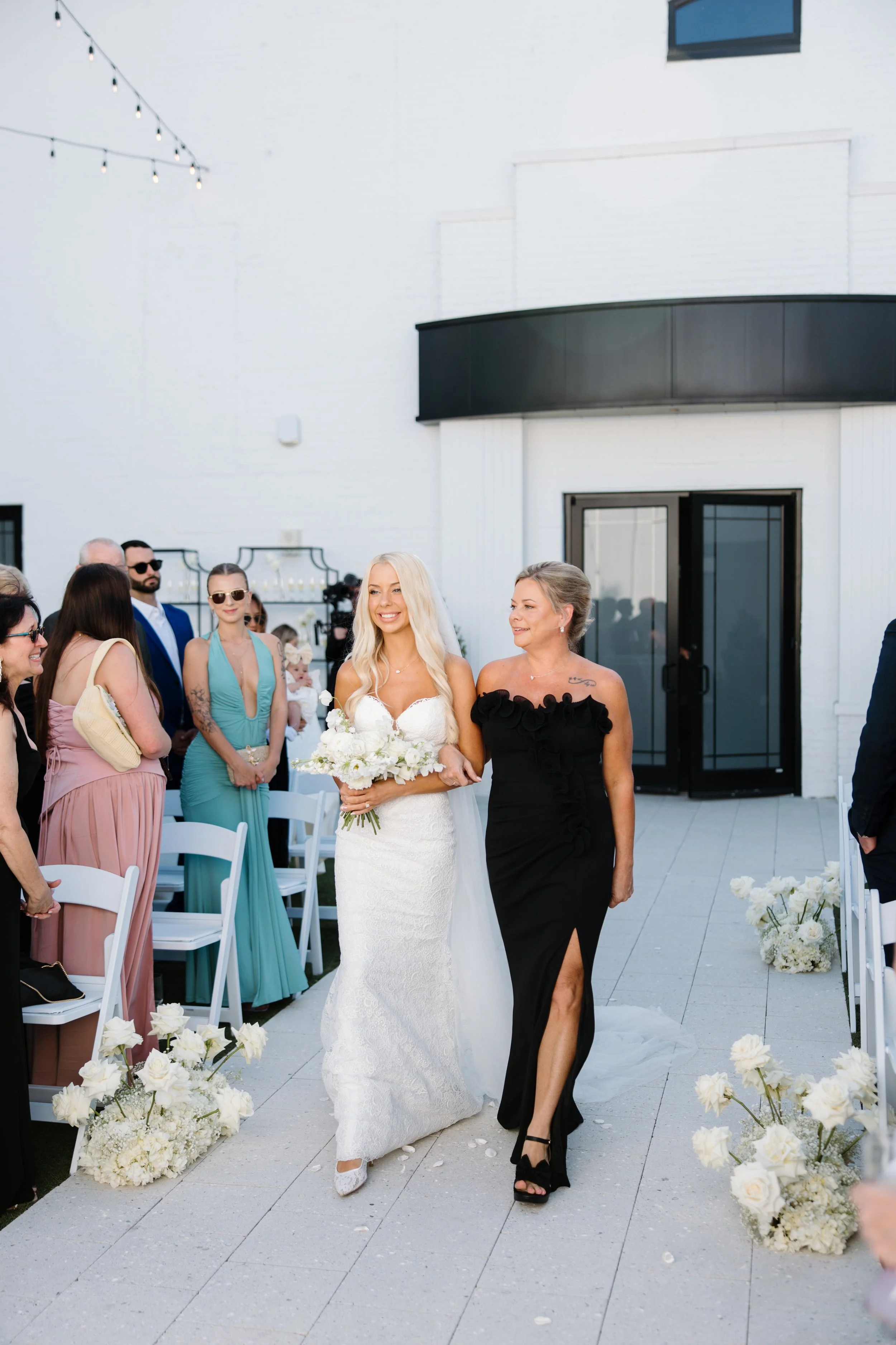 A bride in a white lace wedding dress walking down an aisle, accompanied by a woman in a black dress, at an outdoor wedding ceremony decorated with white flowers. Guests are seated on both sides, and some are standing and watching.