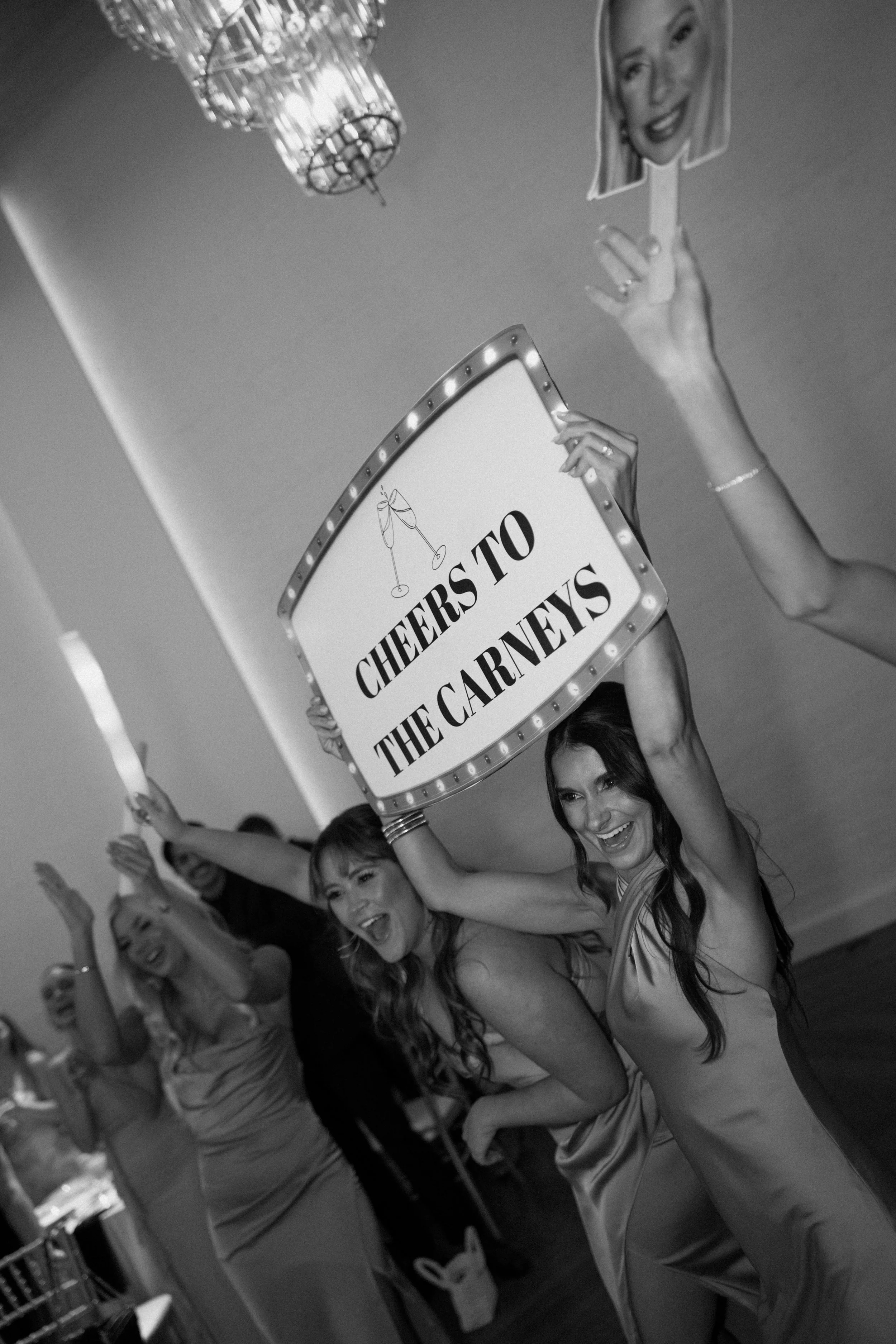 A group of women celebrating and waving their hands in the air, holding a sign that reads "Cheers to the Carneys." They are smiling and appear to be at a party or celebration.