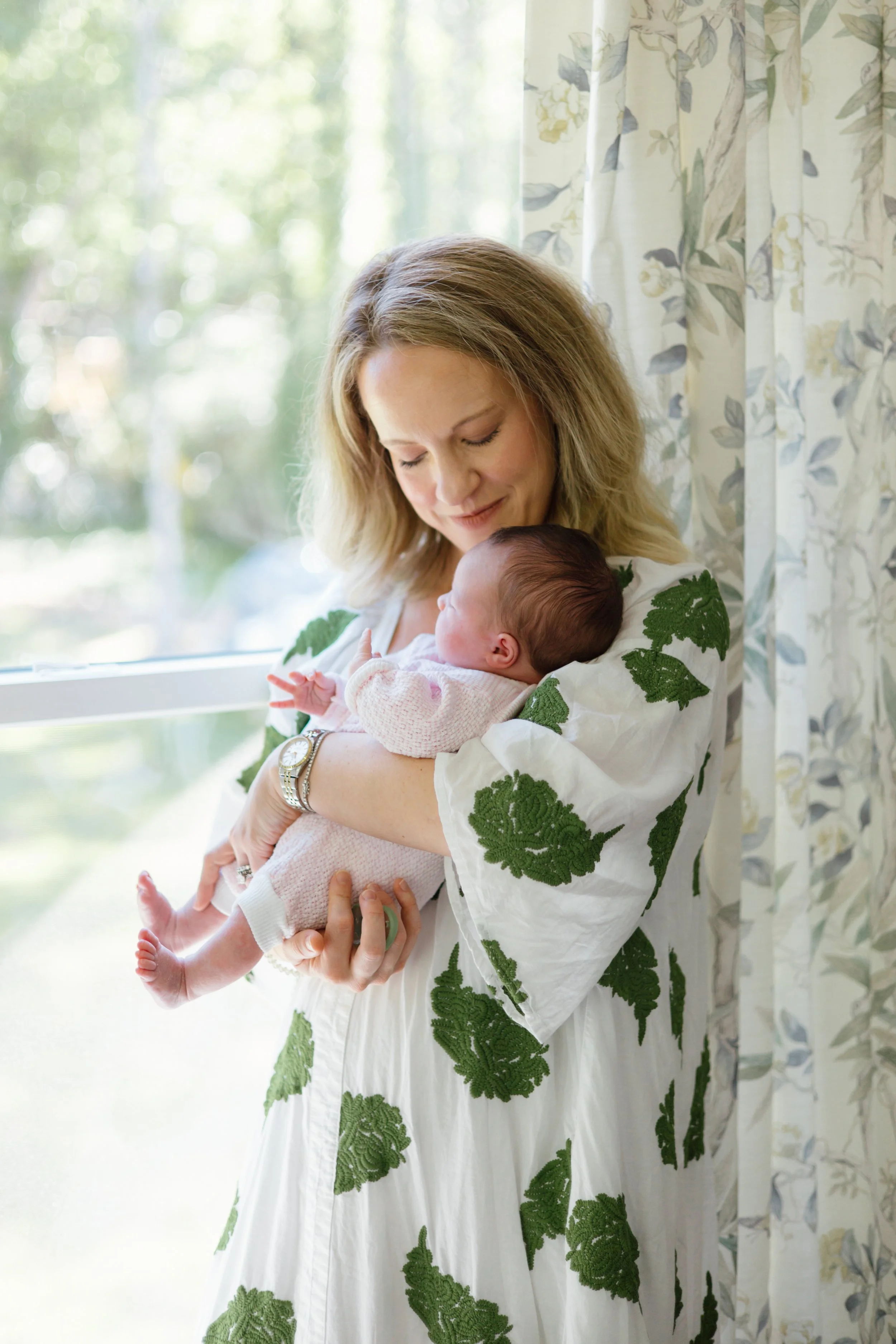 A woman holding a baby near a window with floral curtains, with trees and greenery visible outside.