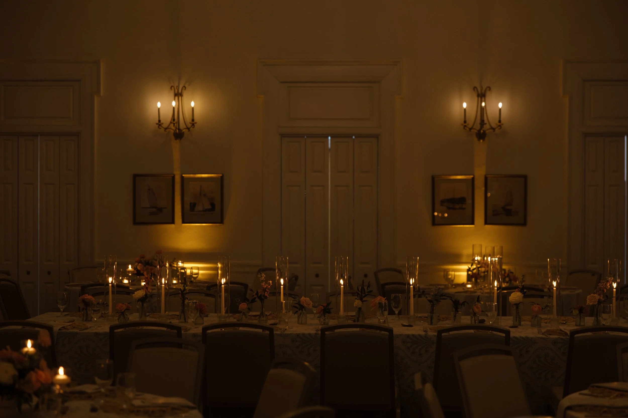 A dimly lit banquet hall with elegant wall sconces, framed sailboat artwork, round tables decorated with floral centerpieces and candles for a formal event.