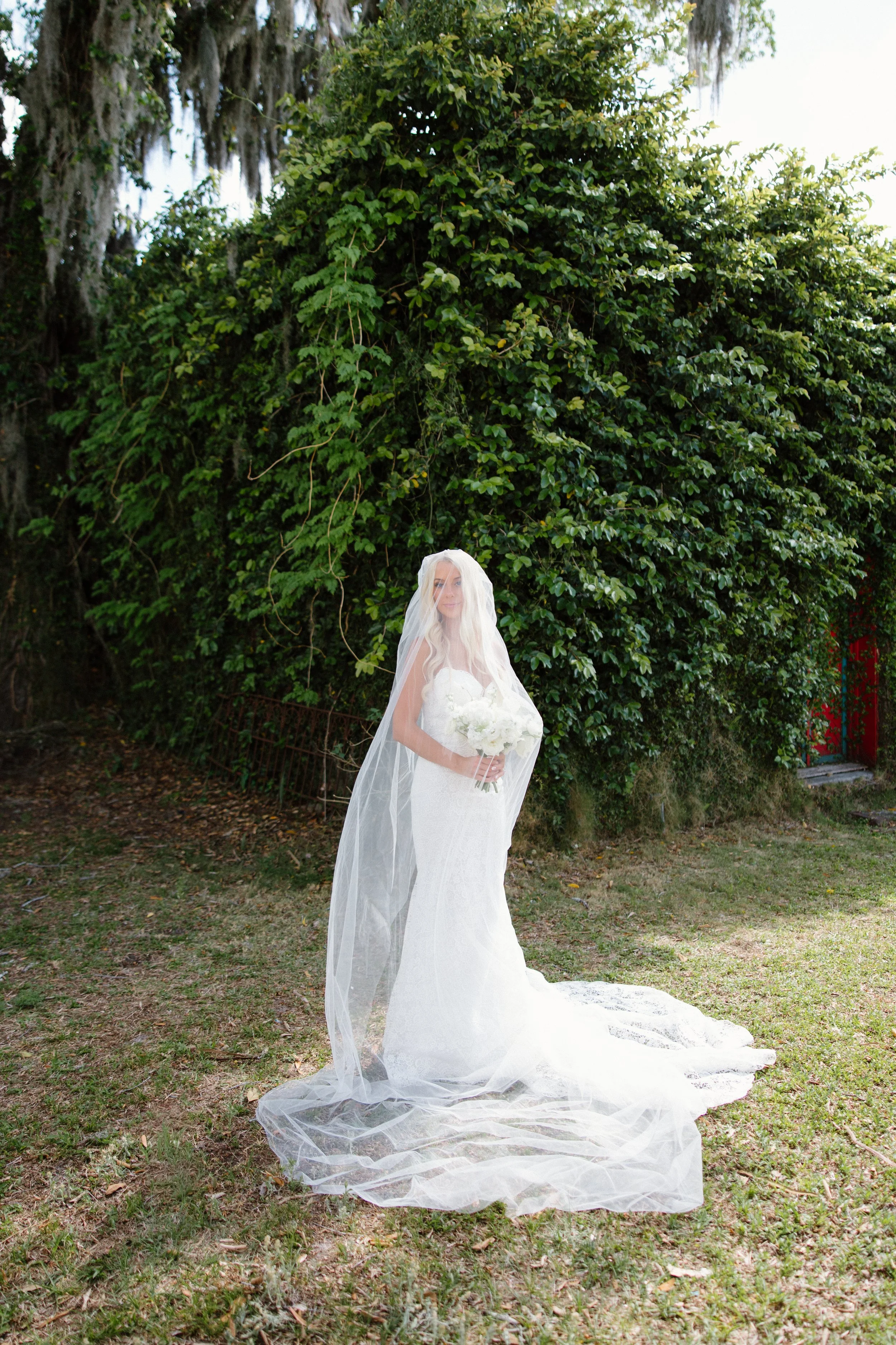 A bride in a white wedding dress and veil holding a bouquet of white flowers, standing outdoors on a grassy area with lush green trees in the background.