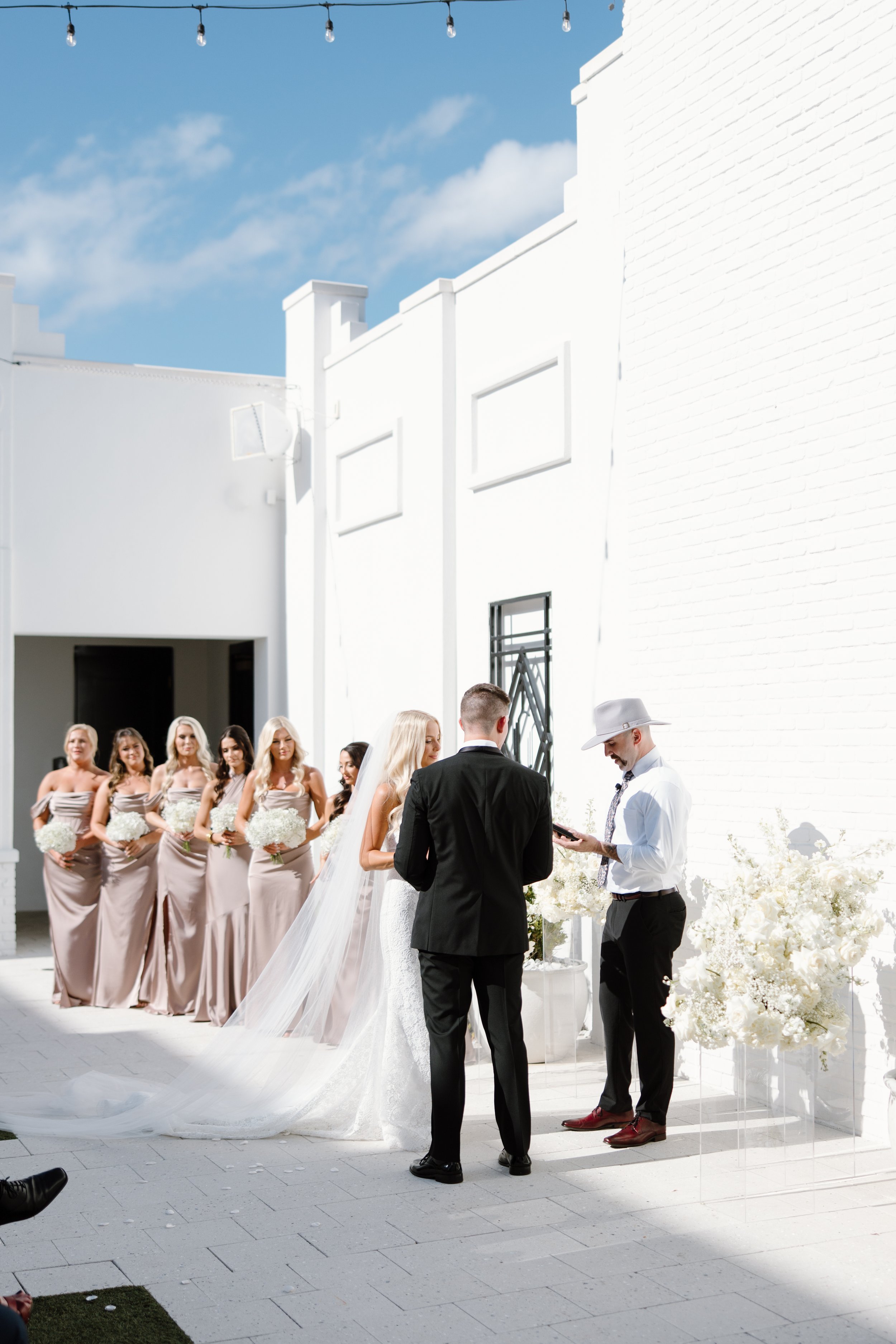A wedding ceremony outdoors with a bride and groom standing in front of an officiant. Bridesmaids in beige dresses holding white bouquets stand behind them, with a white brick building and string lights overhead. The sky is blue with some clouds.