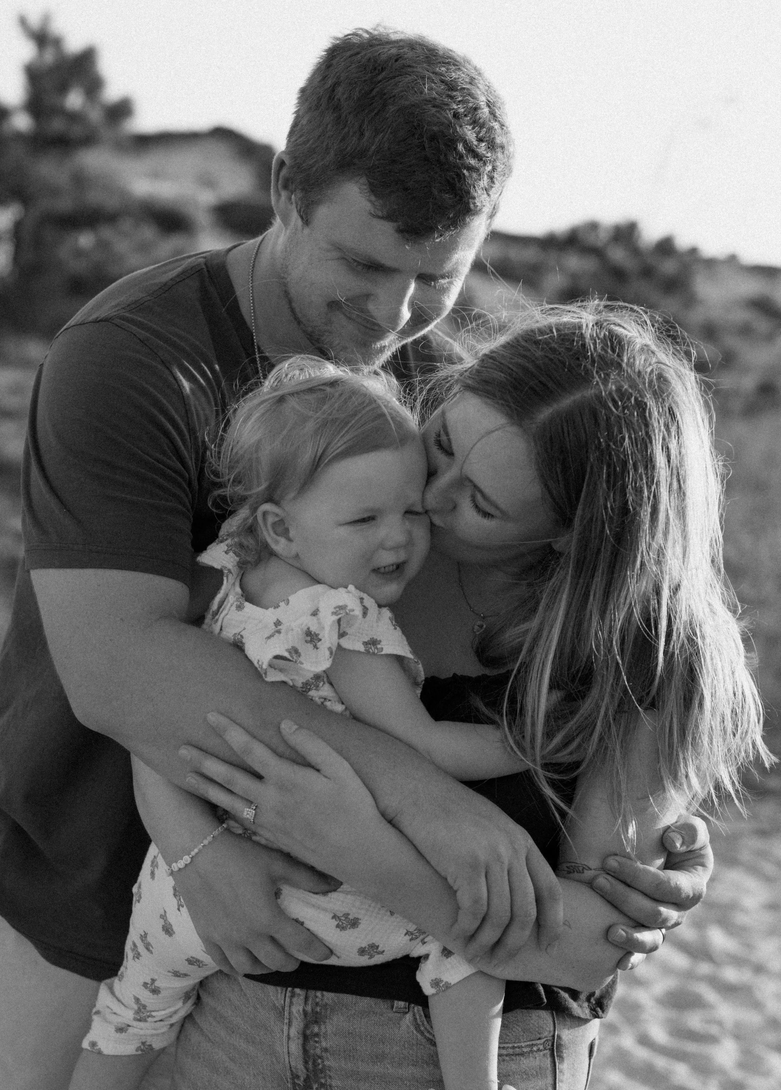 A black and white photograph of a family at the beach showing a woman, a man, and a young girl. The woman is kissing the girl on the head, and the man is smiling with his head bowed down as he embraces them. The background includes a sandy beach and some rocks.
