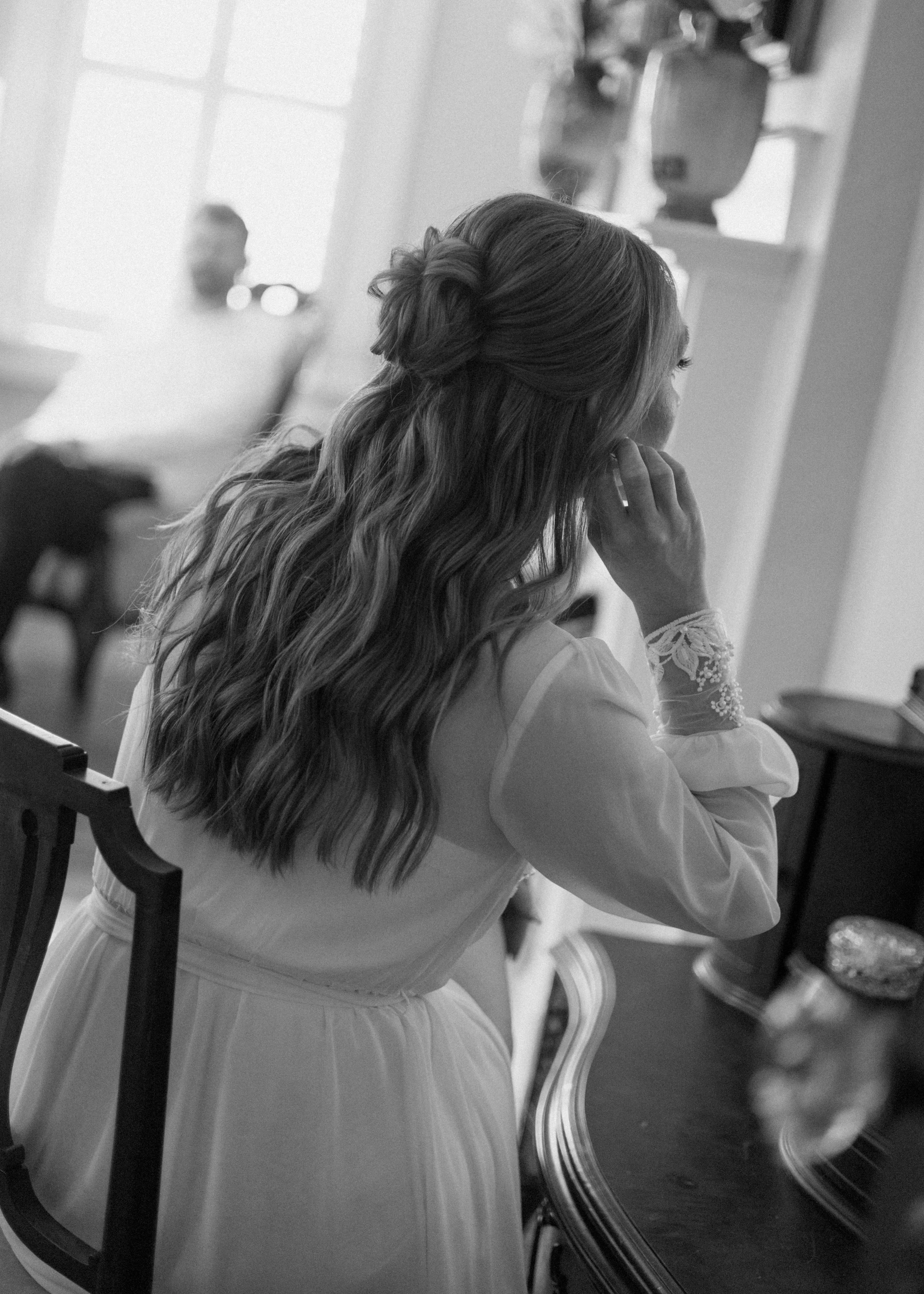A woman with long, wavy hair styled in an elegant updo seated at a vanity or dresser. She is wearing a white, long-sleeved dress with lace detail on the sleeves. The woman is resting her chin on her hand, with a contemplative or thoughtful expression. The background features a large window with daylight streaming in, and reflective surfaces and decorative objects are visible, creating a bright, serene atmosphere.