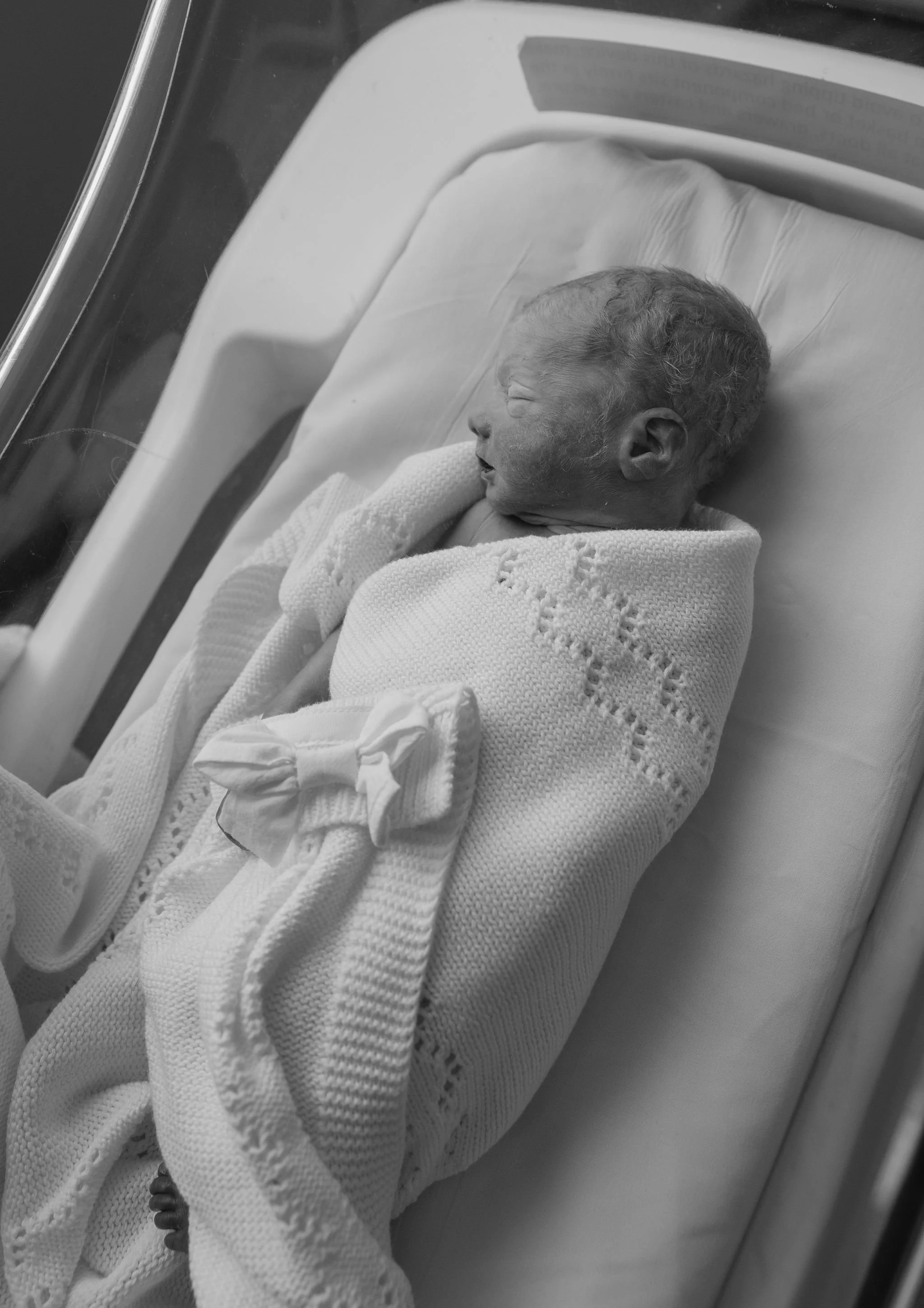Black and white photo of a newborn baby sleeping in an incubator or hospital bassinet, wrapped in a knitted blanket.