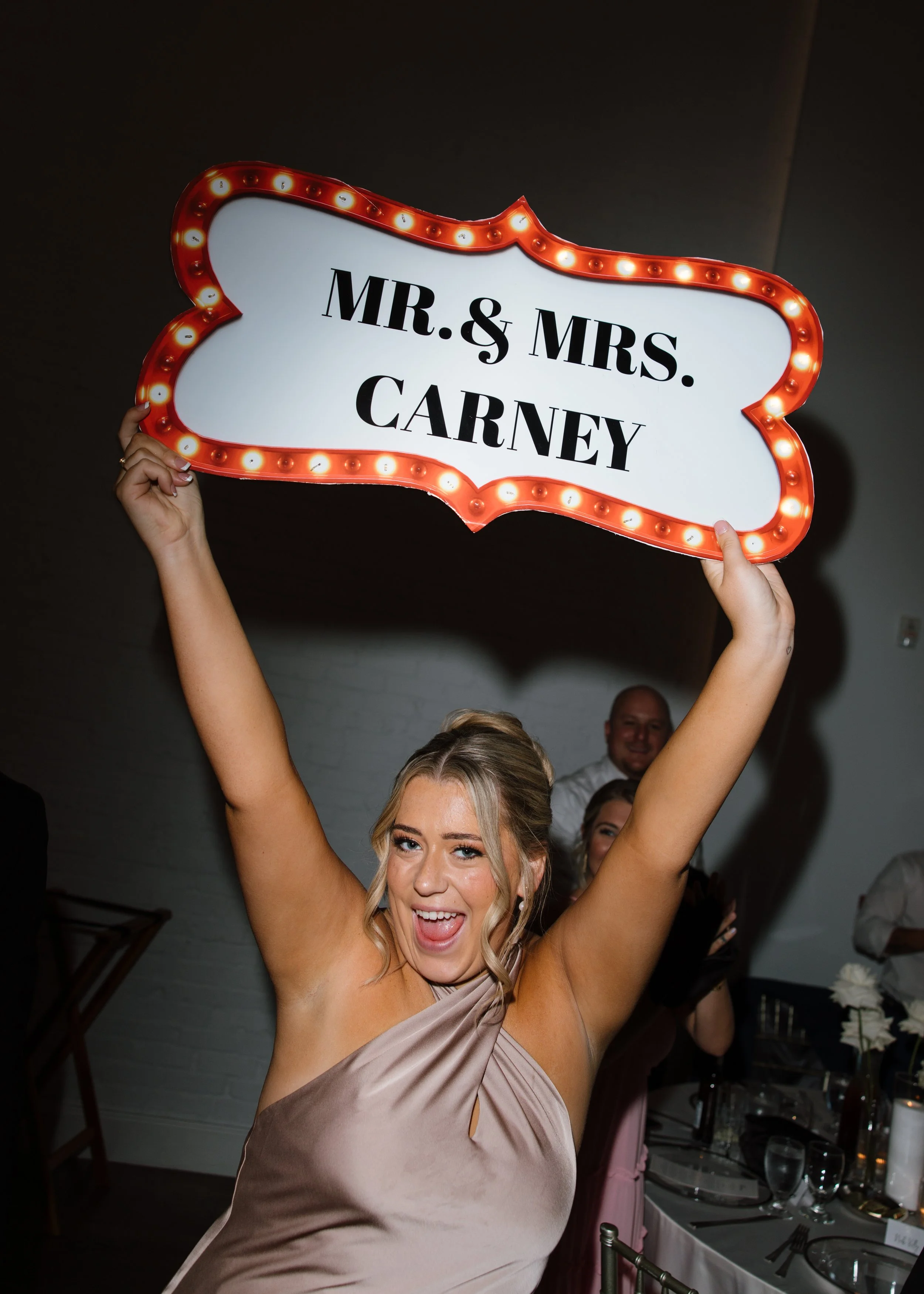 A woman in a beige dress celebrating at a wedding reception, holding a decorative sign that says 'MR. & MRS. CARNEY' with lightbulbs around the border, smiling and appearing joyful.