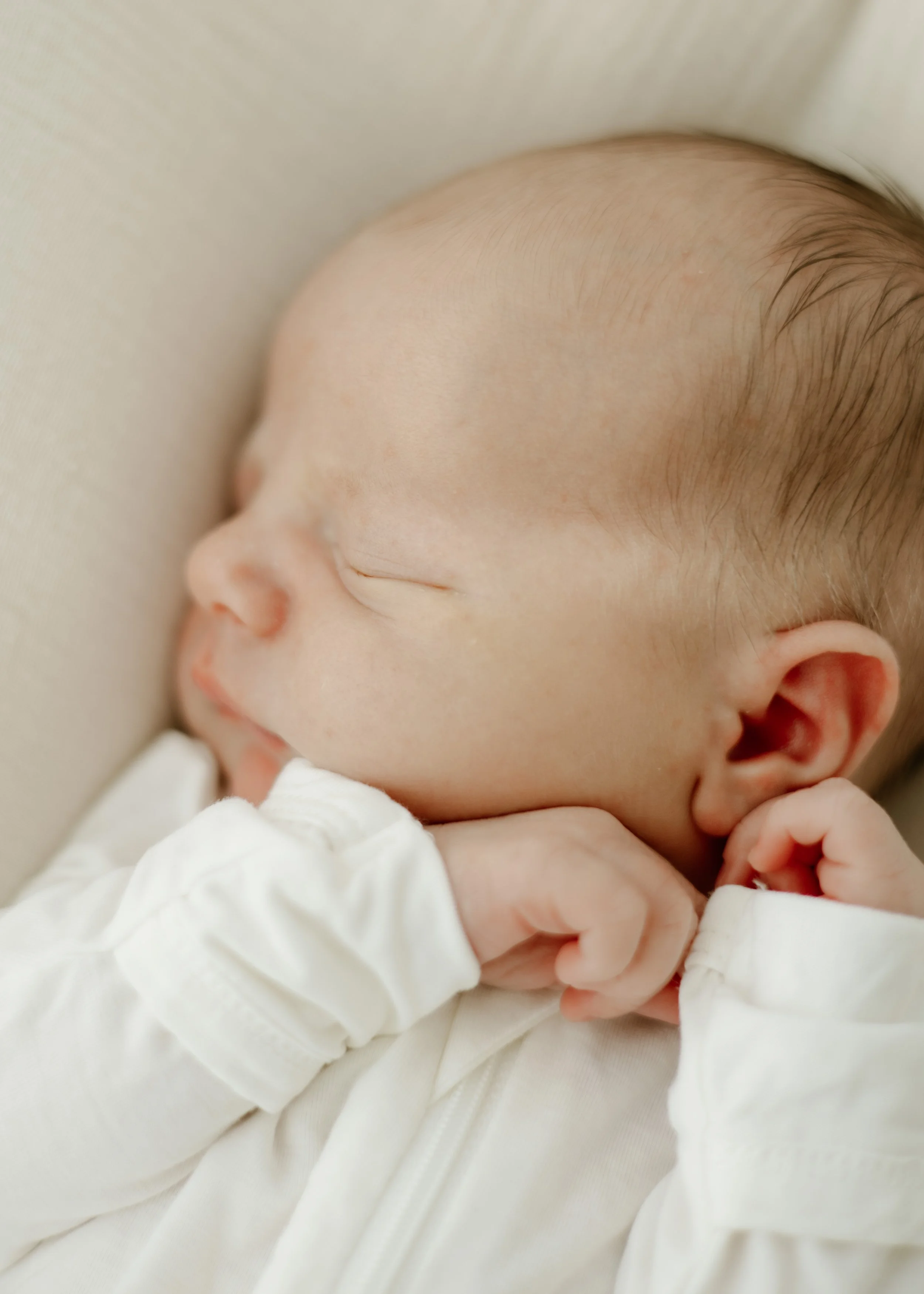 Close-up of a sleeping baby with delicate features, wearing a white outfit, lying on a soft surface.