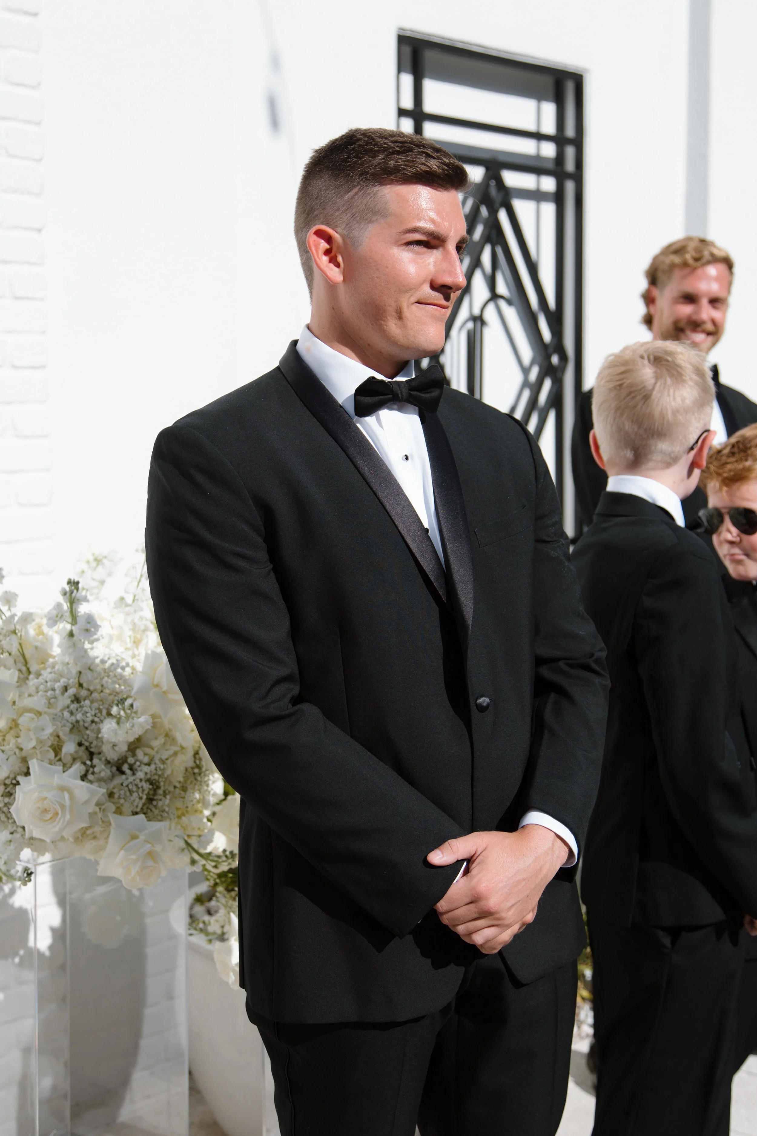 Young man in a tuxedo standing with arms crossed at a wedding ceremony, with others dressed similarly and white floral arrangements in the background.