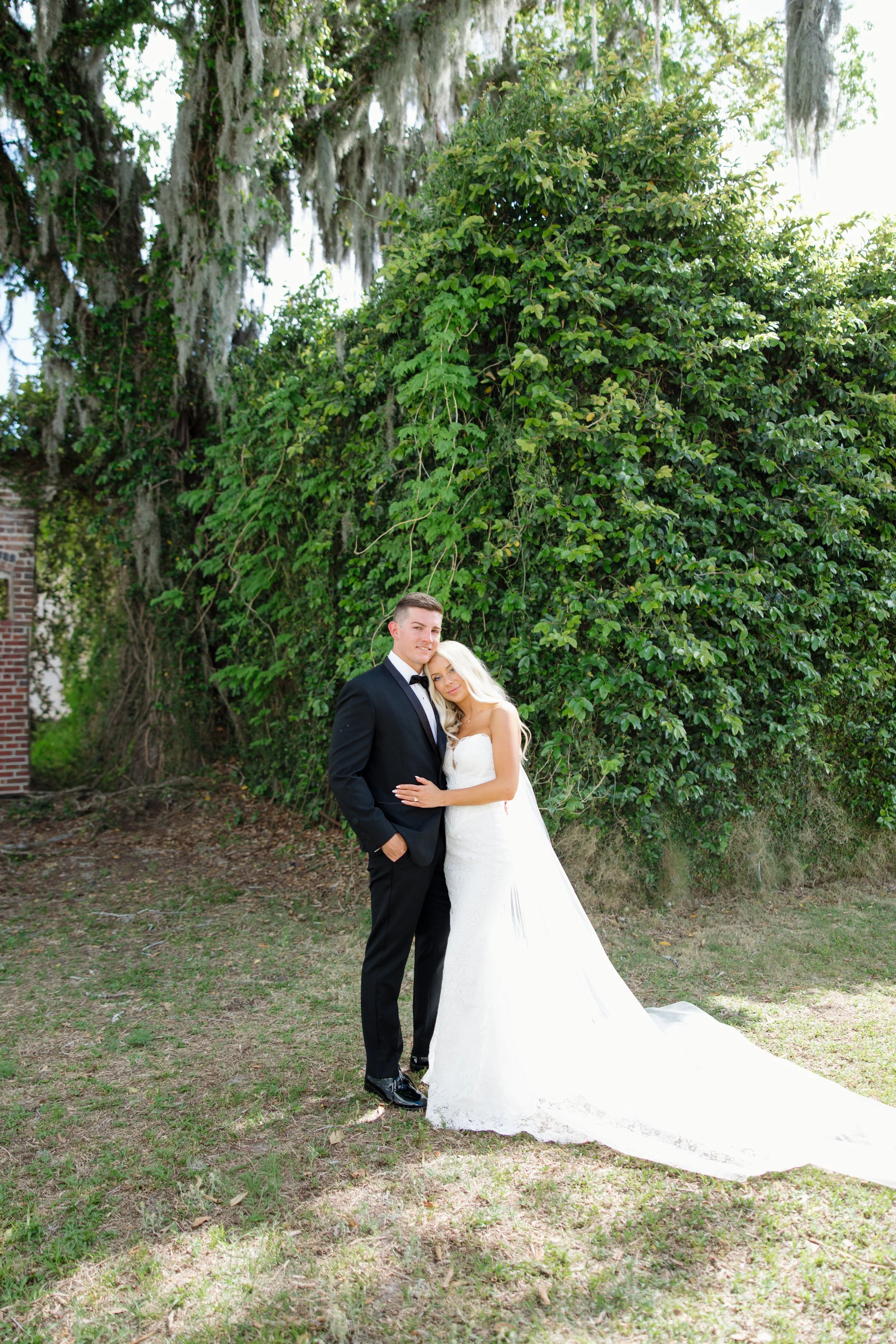 A bride and groom standing close together outdoors, surrounded by greenery and trees, during daytime.