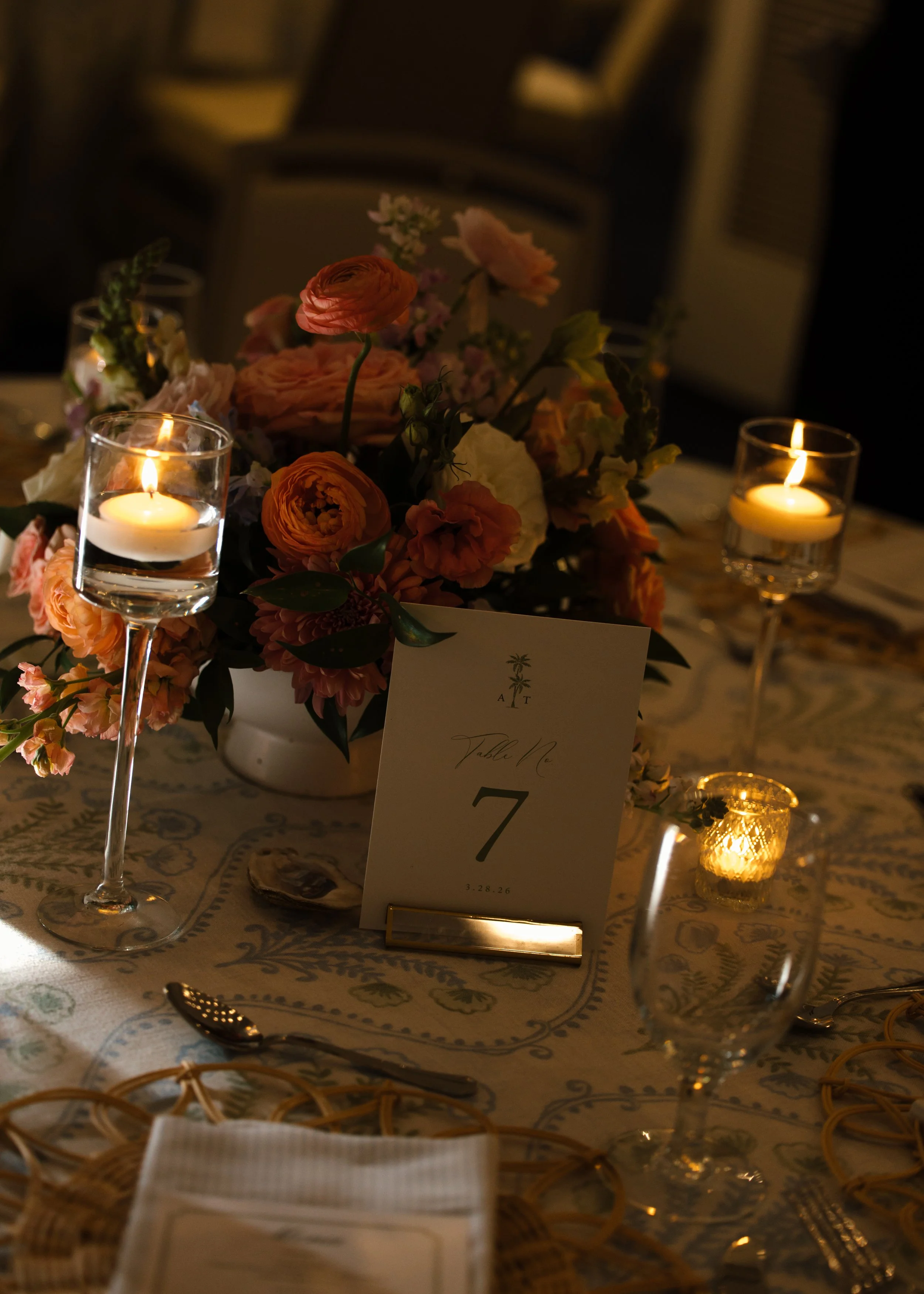 A formal dinner table decorated with a floral arrangement of peach and pink roses, with lit candles in glass holders, a table number card displaying '7', and elegant tableware.