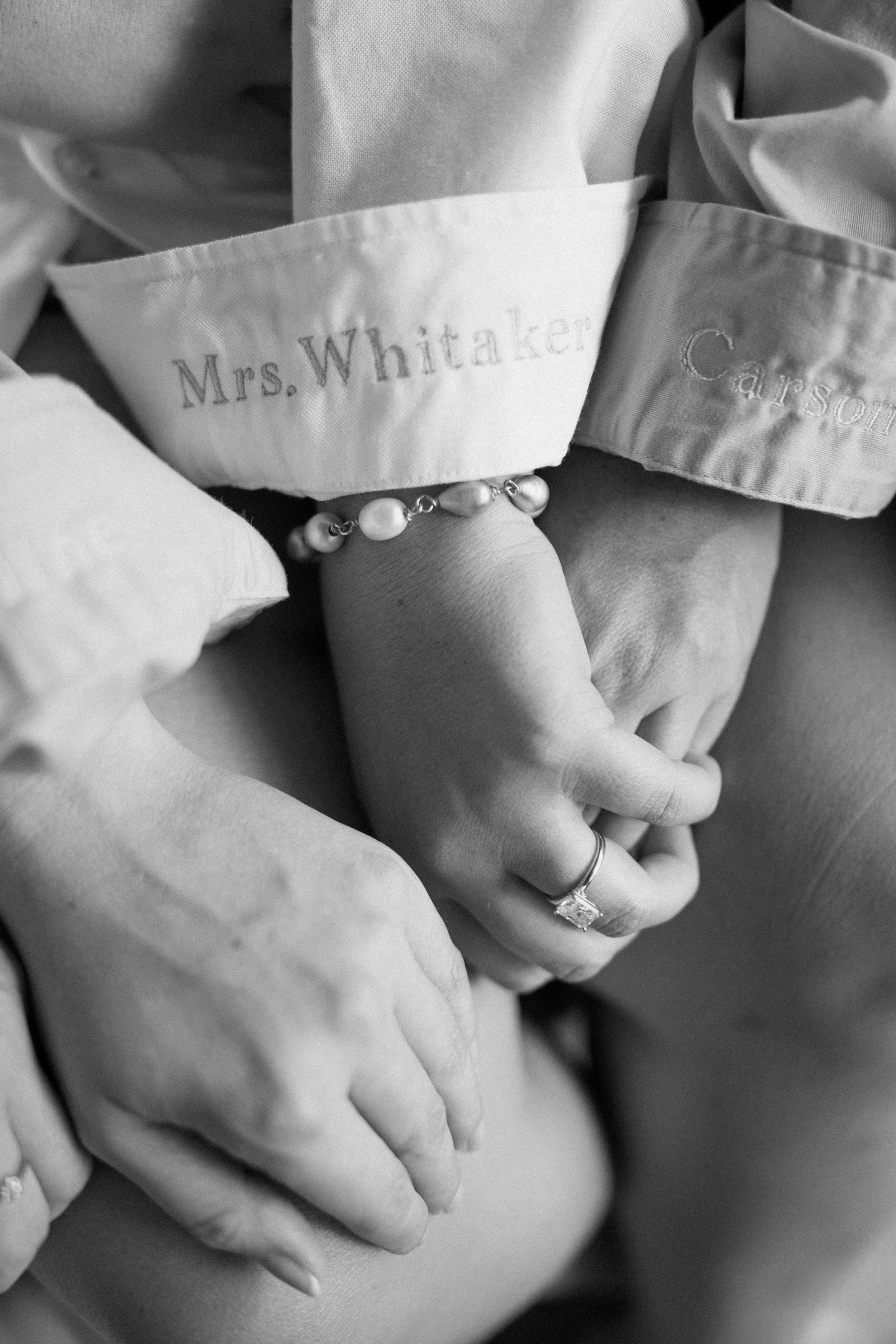 Close-up of hands holding a nurse's or doctor's hand, visible rings on fingers, with a medical bracelet and embroidered name tag reading 'Mrs. Whitaker' on a medical coat.