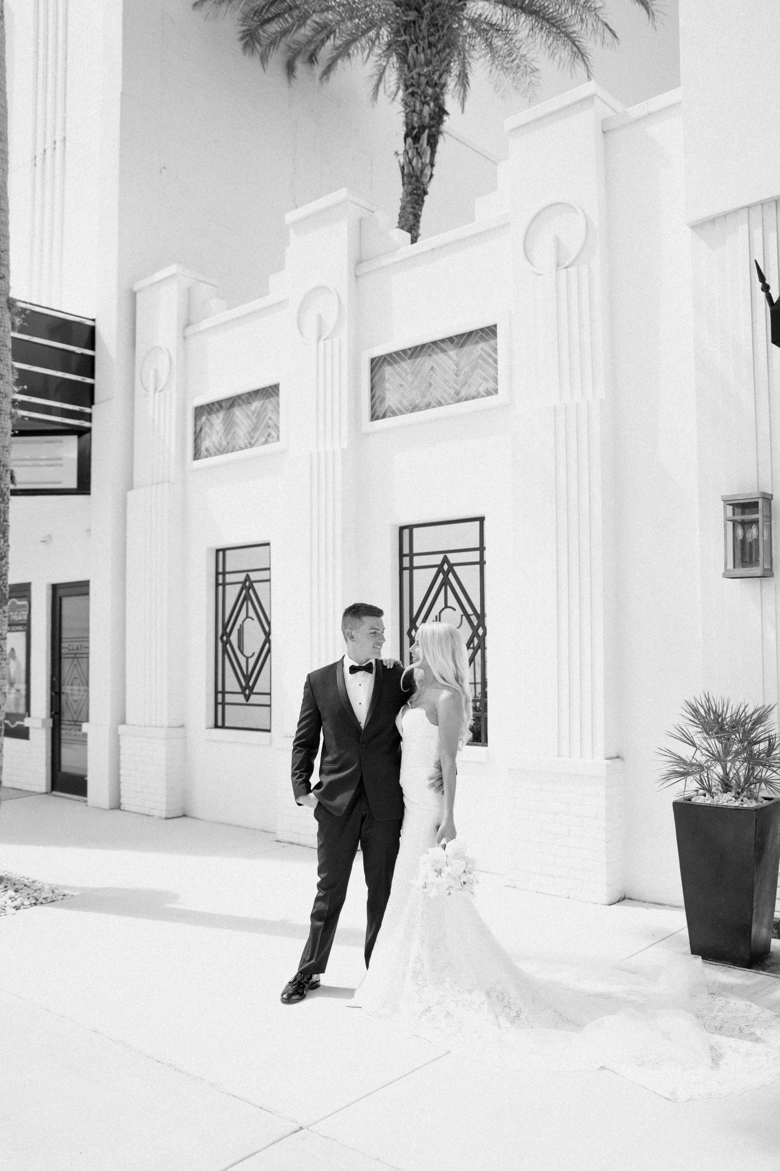 A couple in formal attire, with the man in a tuxedo and the woman in a white dress, standing together outdoors near a white building with palm trees in the background.