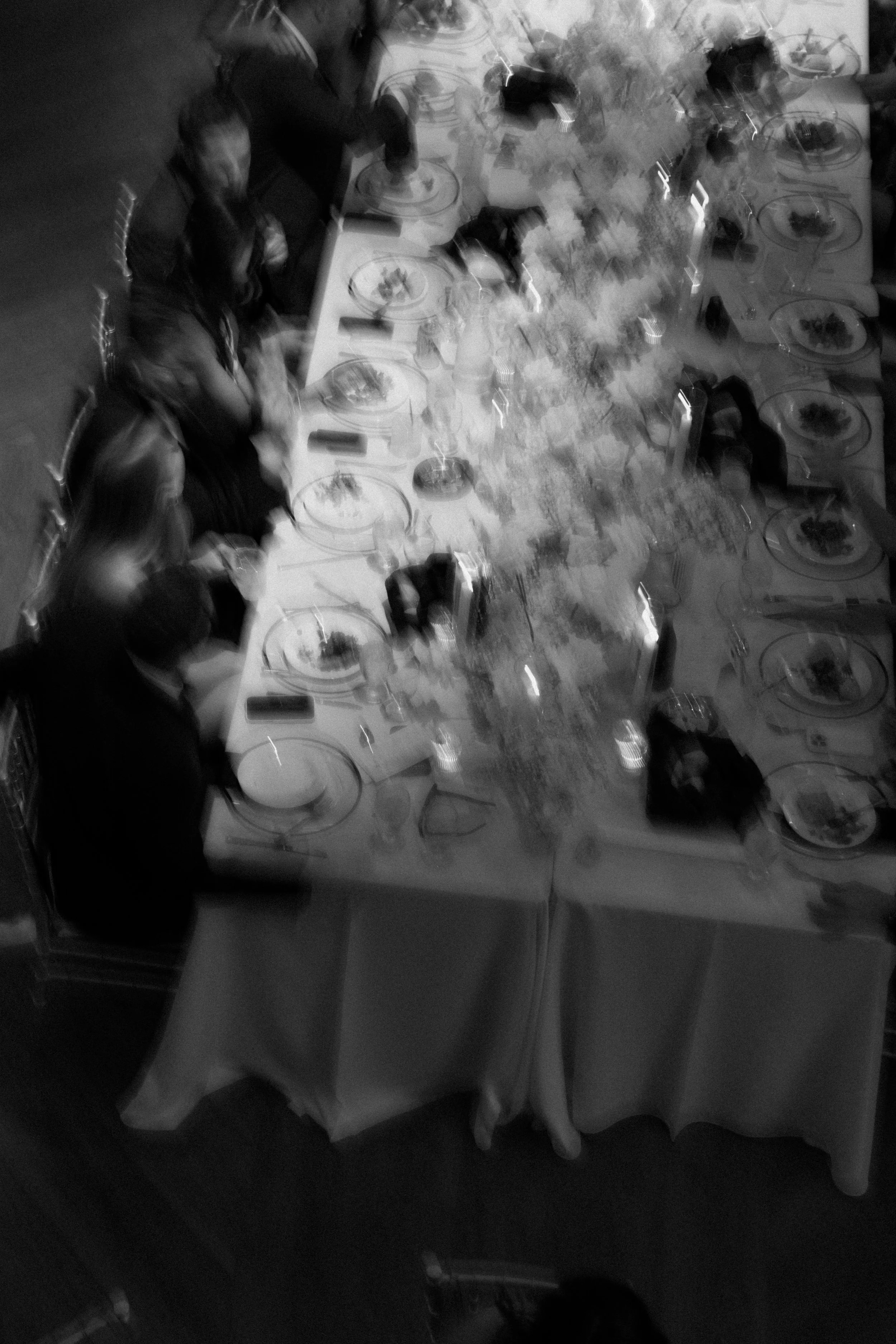 People sitting around a dining table with plates of food, drinks, and floral centerpiece, seen from above. The image is in black and white.