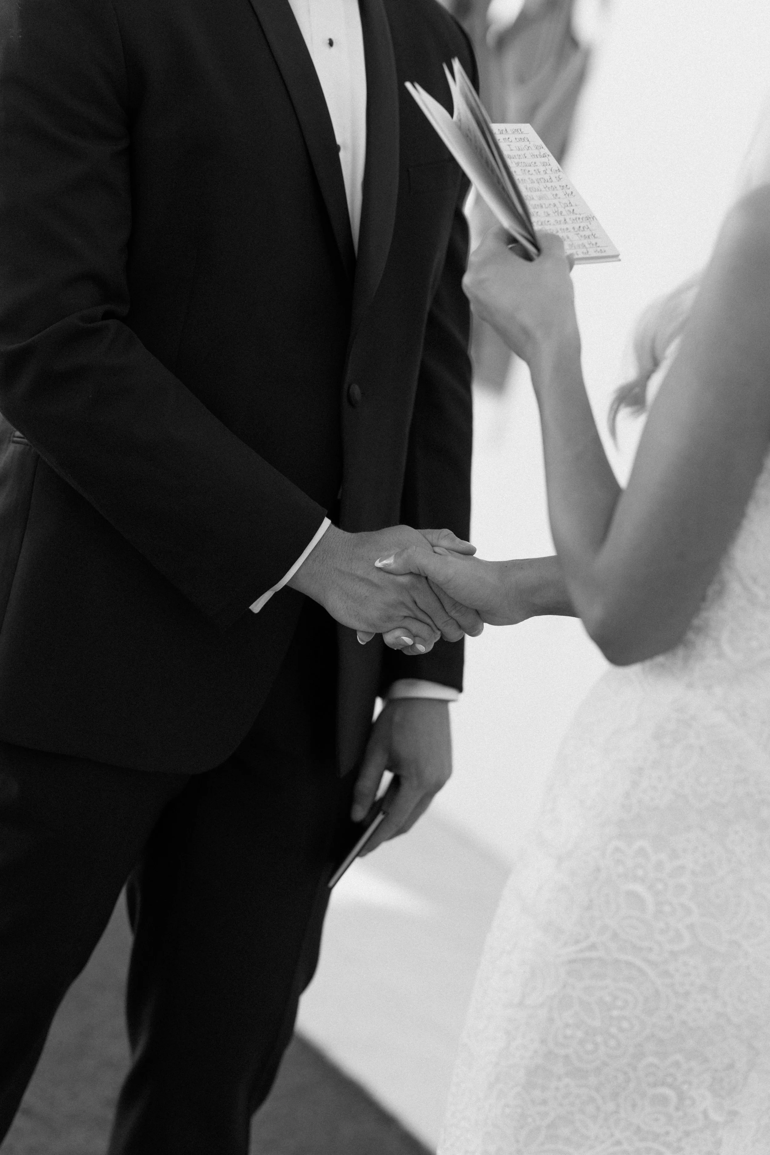 A close-up of a formal wedding ceremony showing a groom in a tuxedo holding hands with a bride in a wedding dress, shaking hands and exchanging vows.
