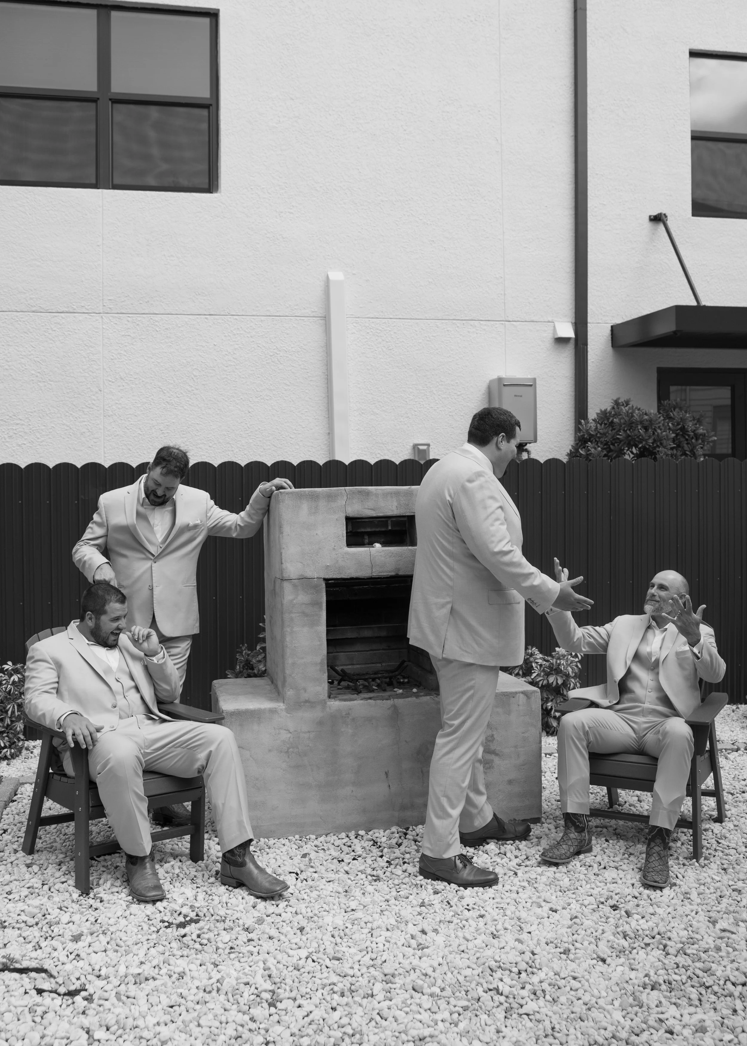 Four men in suits relaxing outdoors around a chimney, two sitting on chairs, one standing, and one gesturing with his hands, on a gravel surface with a building and window in the background.