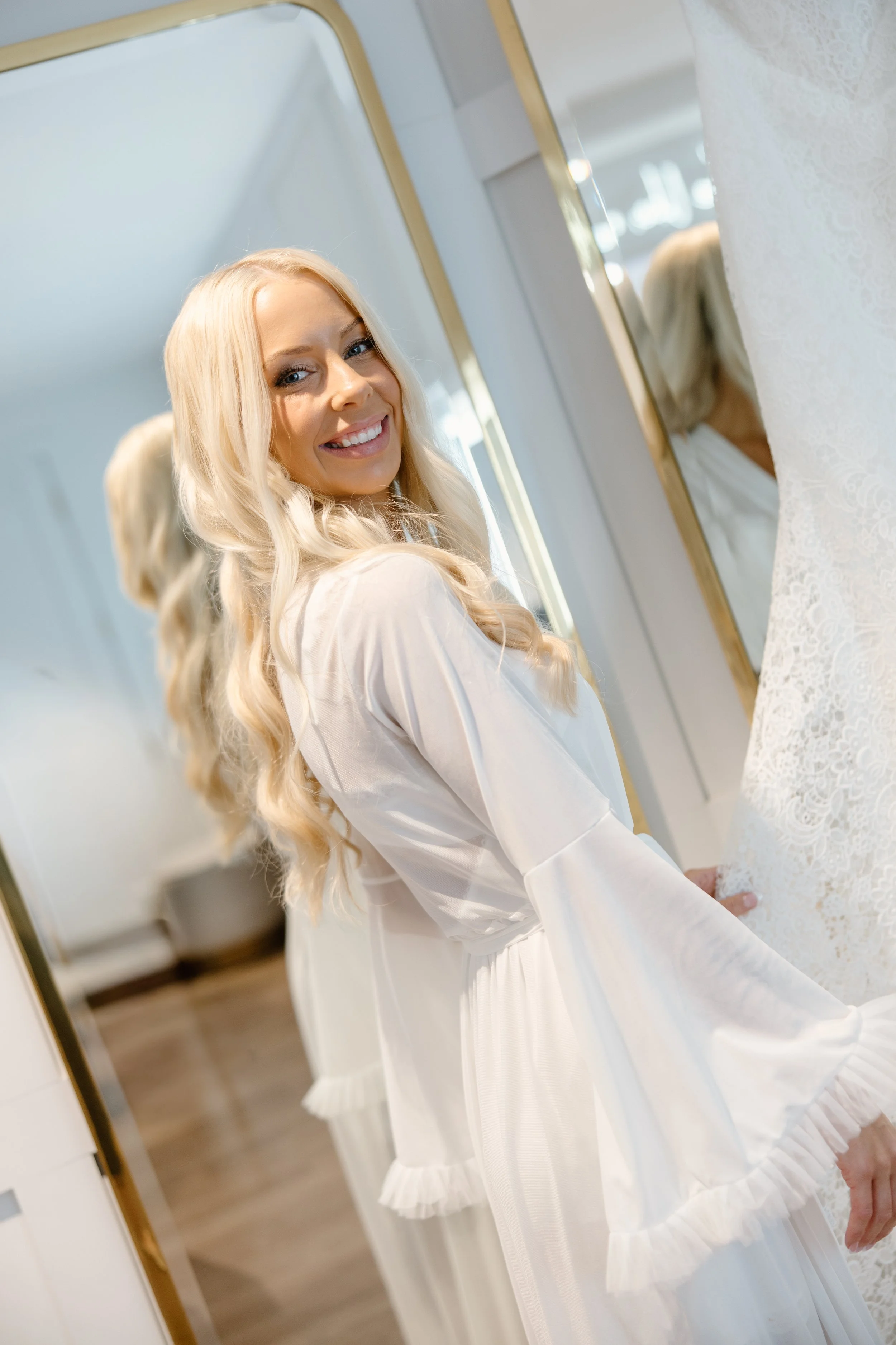 A smiling woman with long, blonde hair wearing a white dress, looking at herself in a mirror.