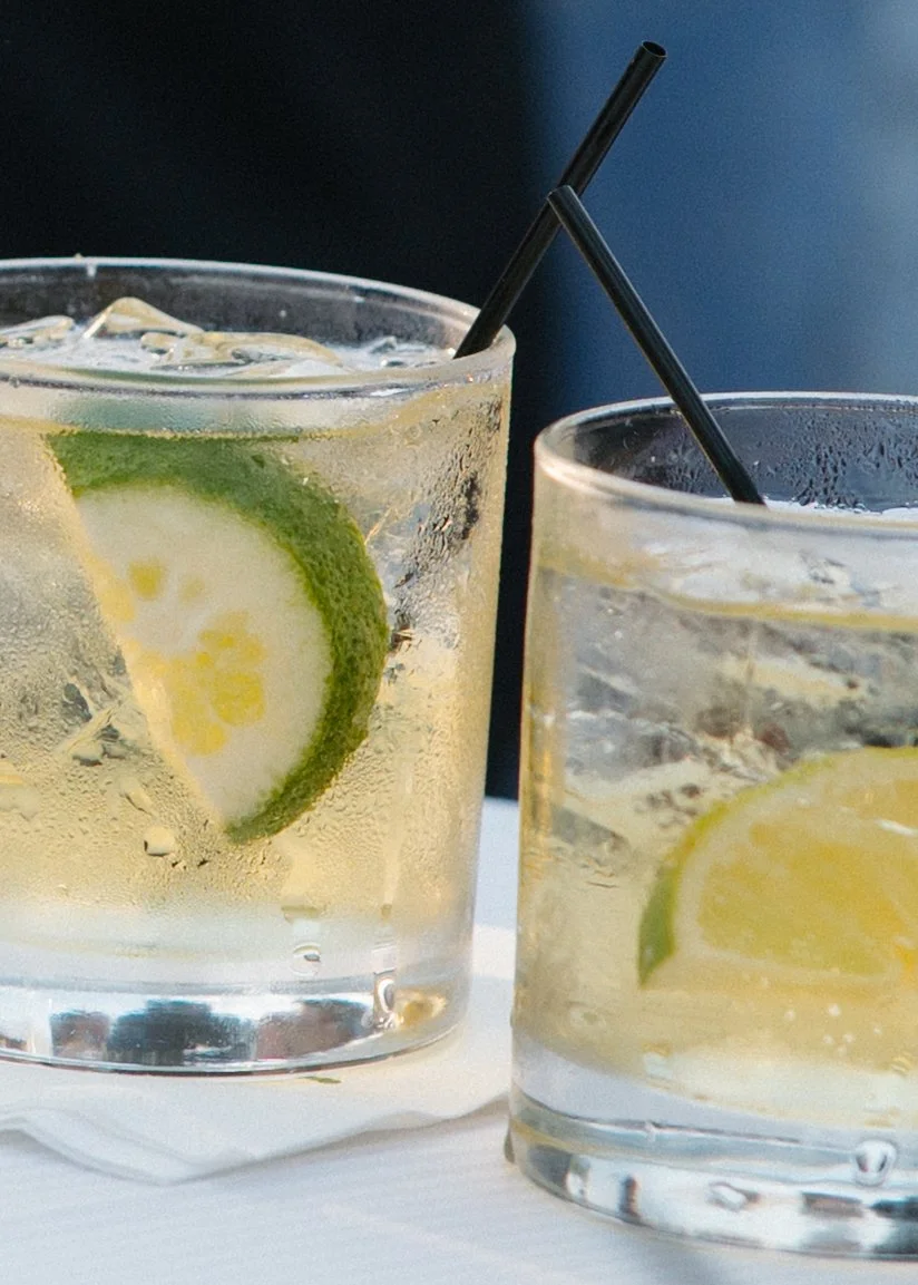 Two glasses of lemon and lime flavored beverages with black straws, ice, and slices of lemon and lime, set on a white surface with a dark background.