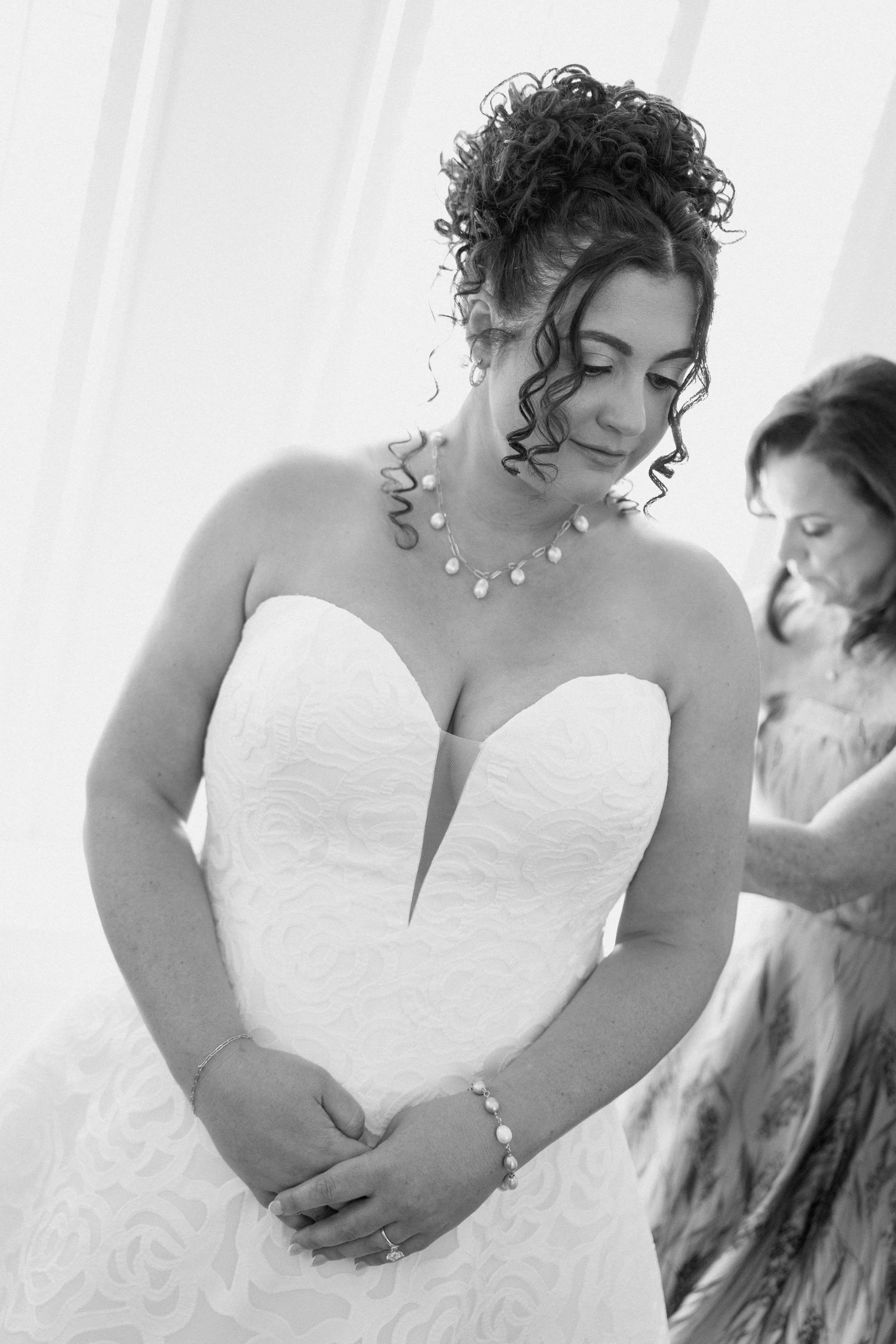 A woman in a strapless white wedding dress with a textured pattern, wearing a pearl necklace, bracelet, and an engagement ring, with another woman in the background wearing a dress with floral patterns.
