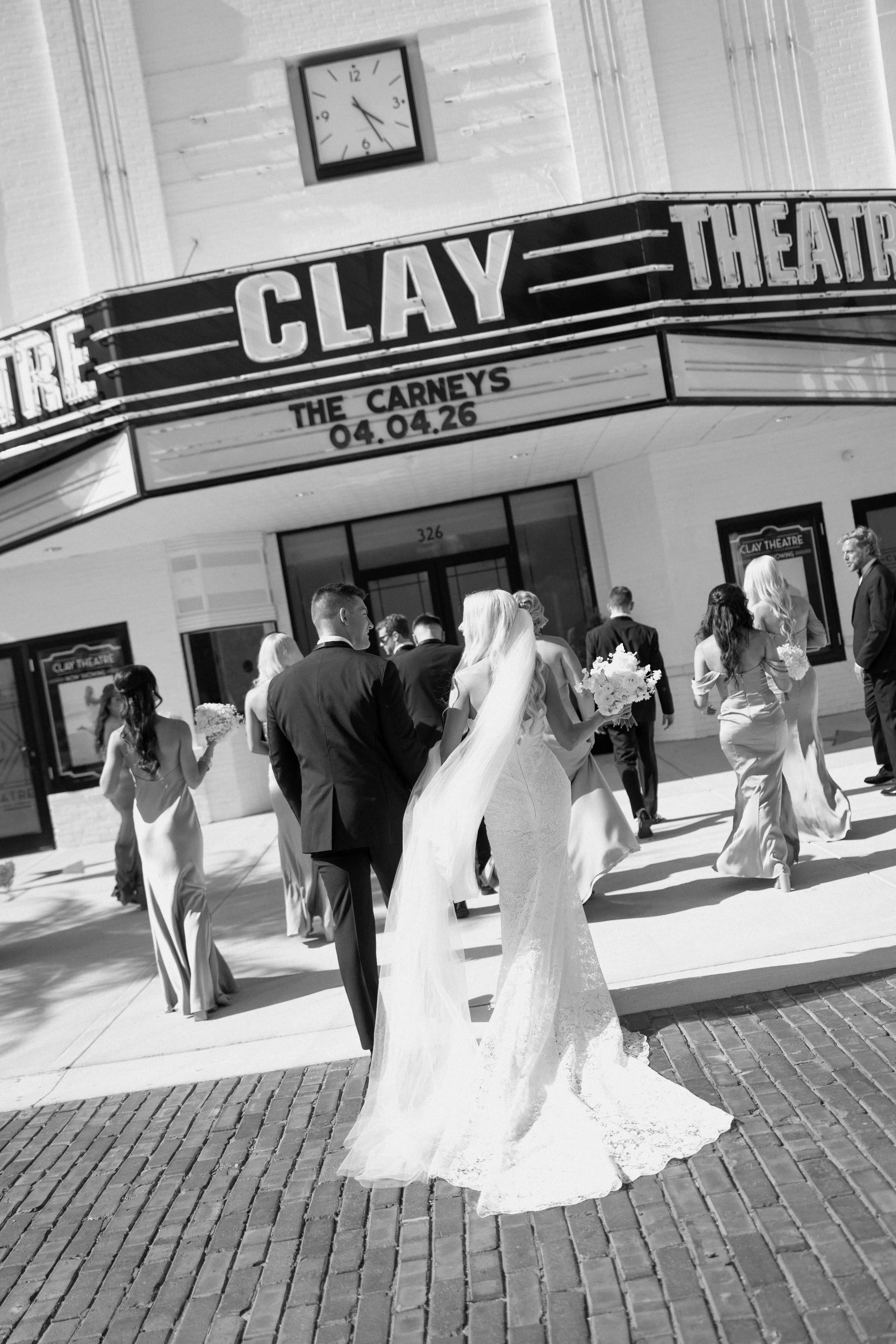 A group of wedding attendees dressed in formal attire, including wedding dresses and suits, walking outside a theater with a marquee sign that reads, 'The Carneys,' and features the date 04.04.26. A large clock is visible on the wall above the marque