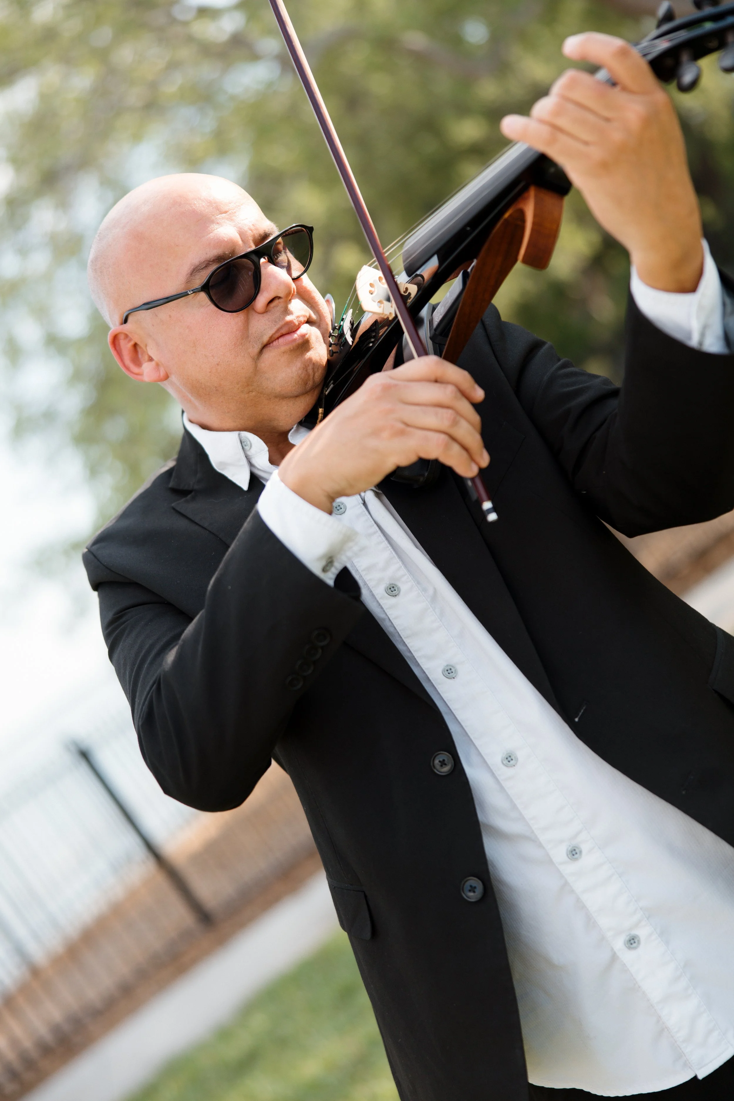 A man with a bald head, sunglasses, and a black suit playing a violin outdoors.