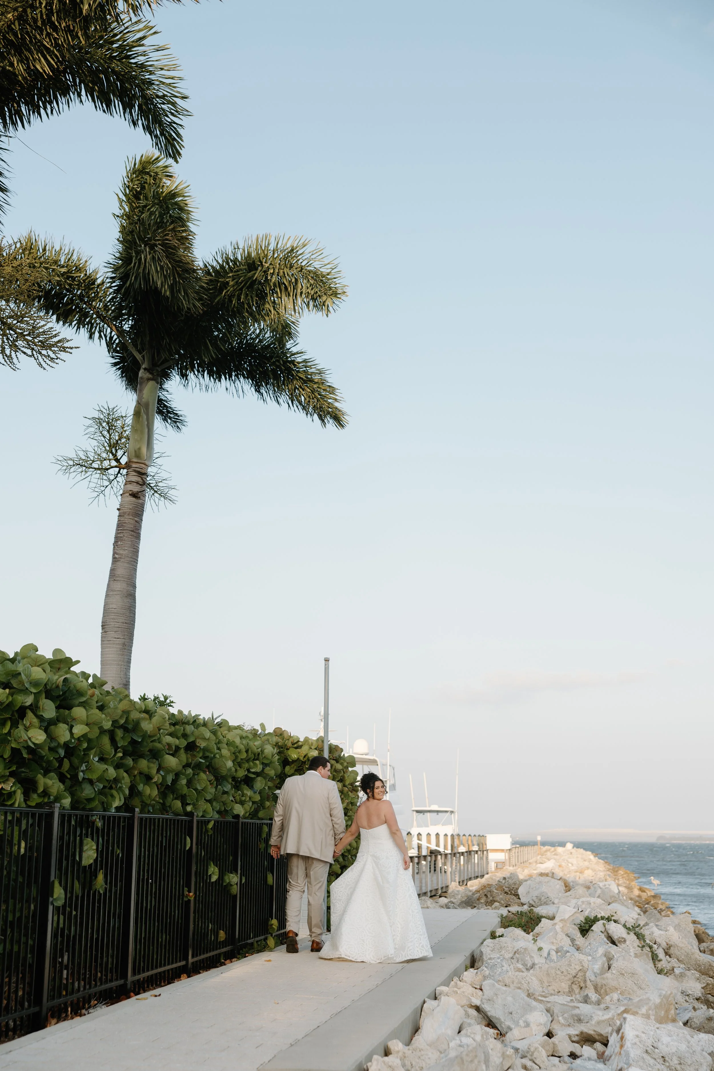 A bride and groom walking hand in hand along a waterfront promenade with palm trees and boats in the background during a wedding photo shoot.