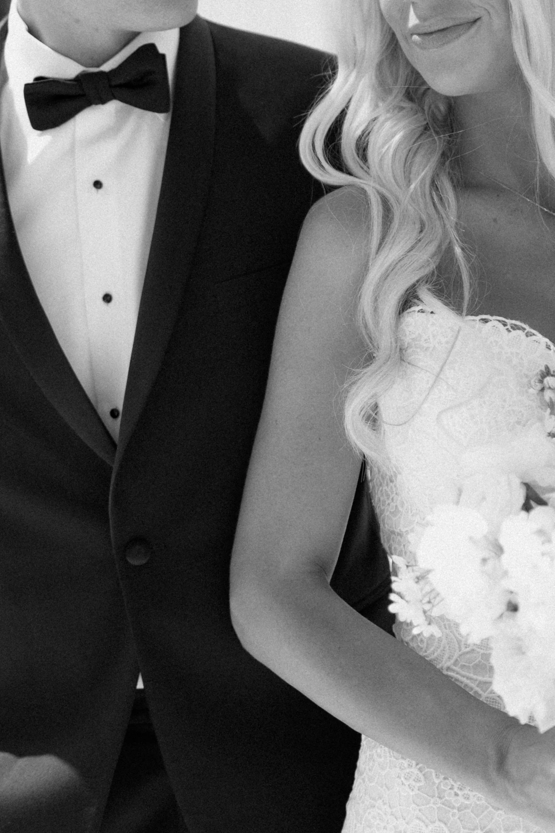 Close-up of a couple dressed in formal attire, with the man wearing a tuxedo and bow tie, and the woman in a lace dress with styled, wavy hair.