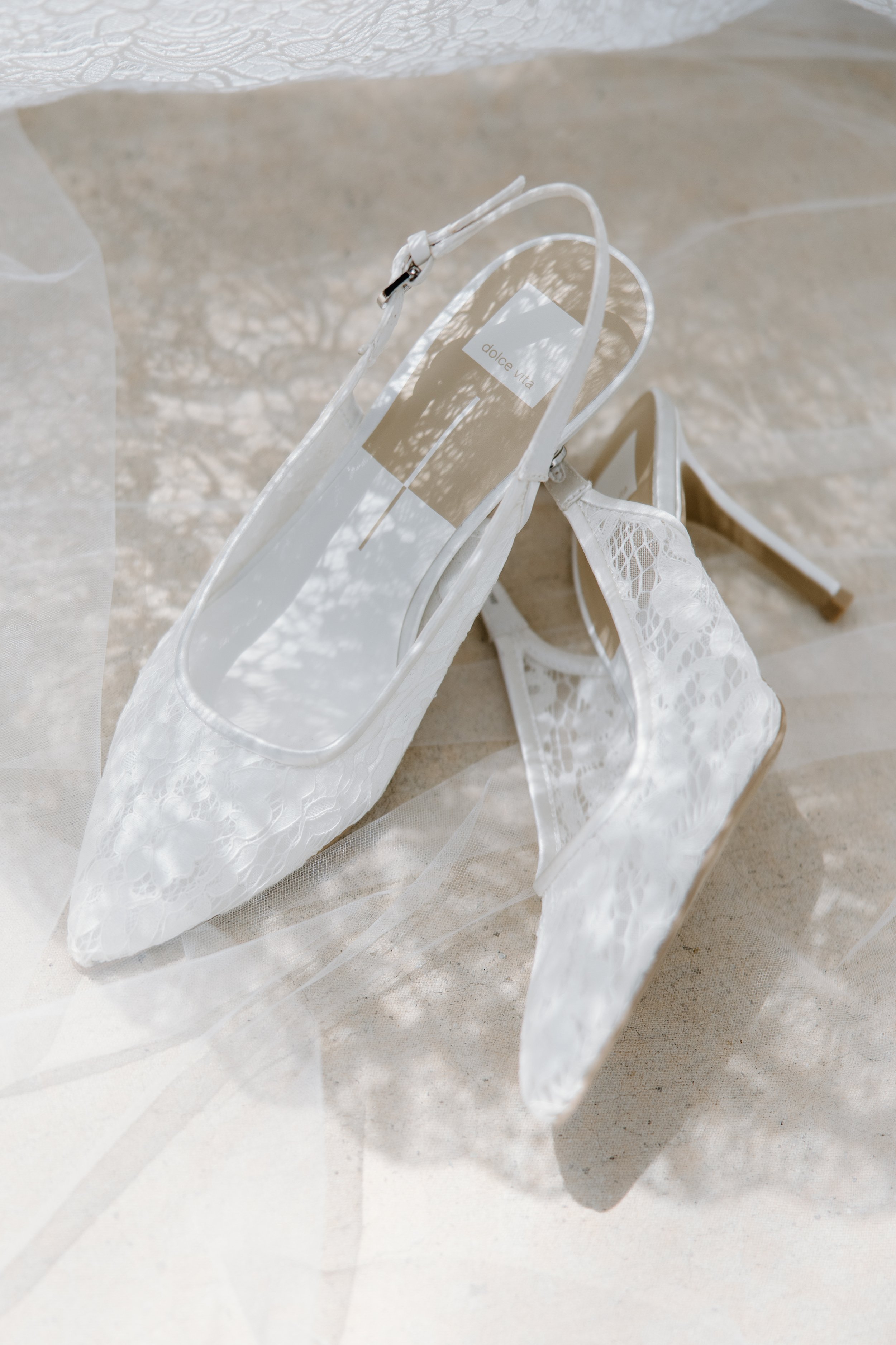 White lace high-heeled shoes with transparent straps, placed on beige surface. A white lace veil is partially visible in the foreground.