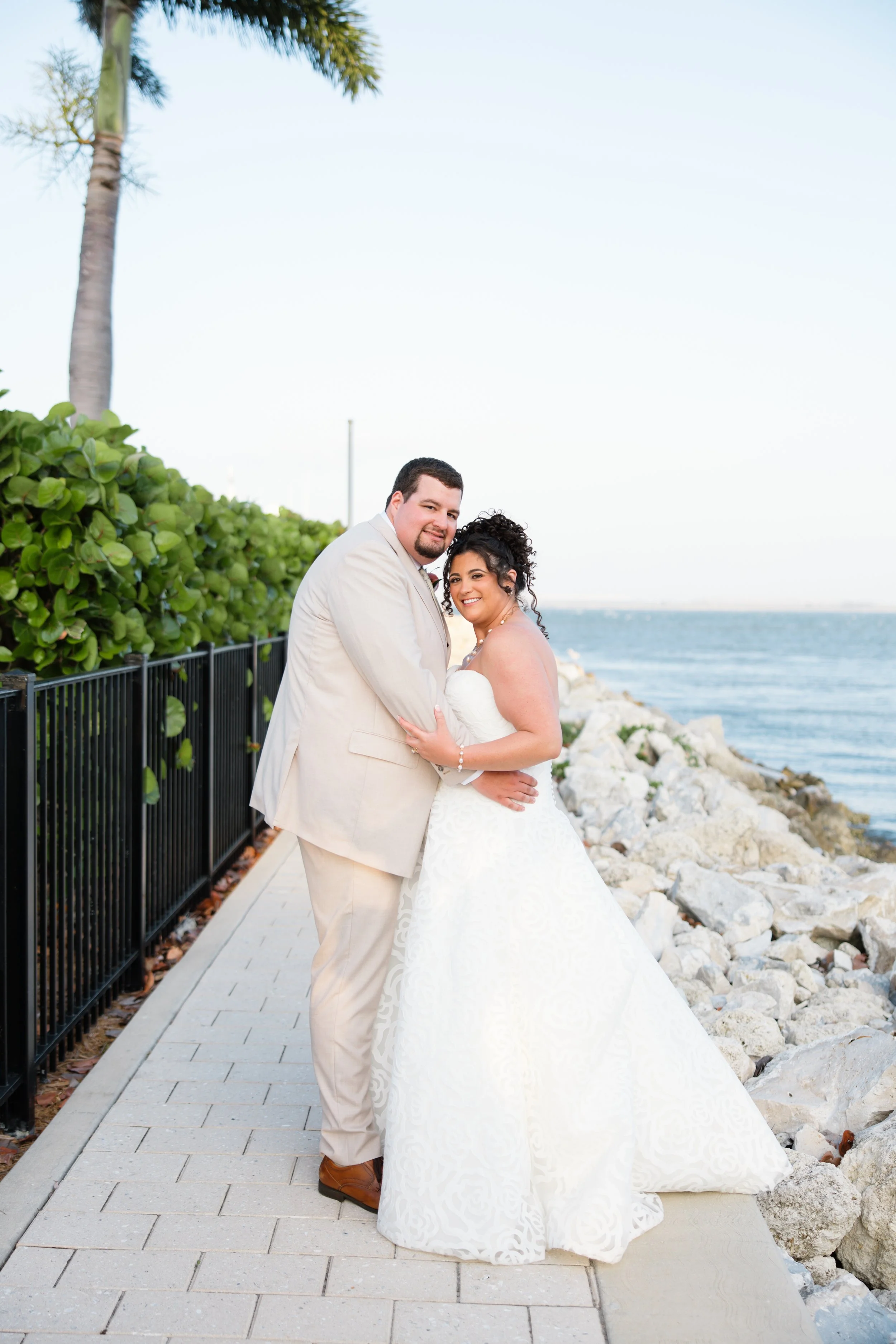 A newlywed couple stands close together on a paved walkway near a rocky shoreline. The groom, wearing a light beige suit, and the bride, in a white lace wedding gown, smile at the camera. Behind them are green bushes, palm trees, and the ocean with a