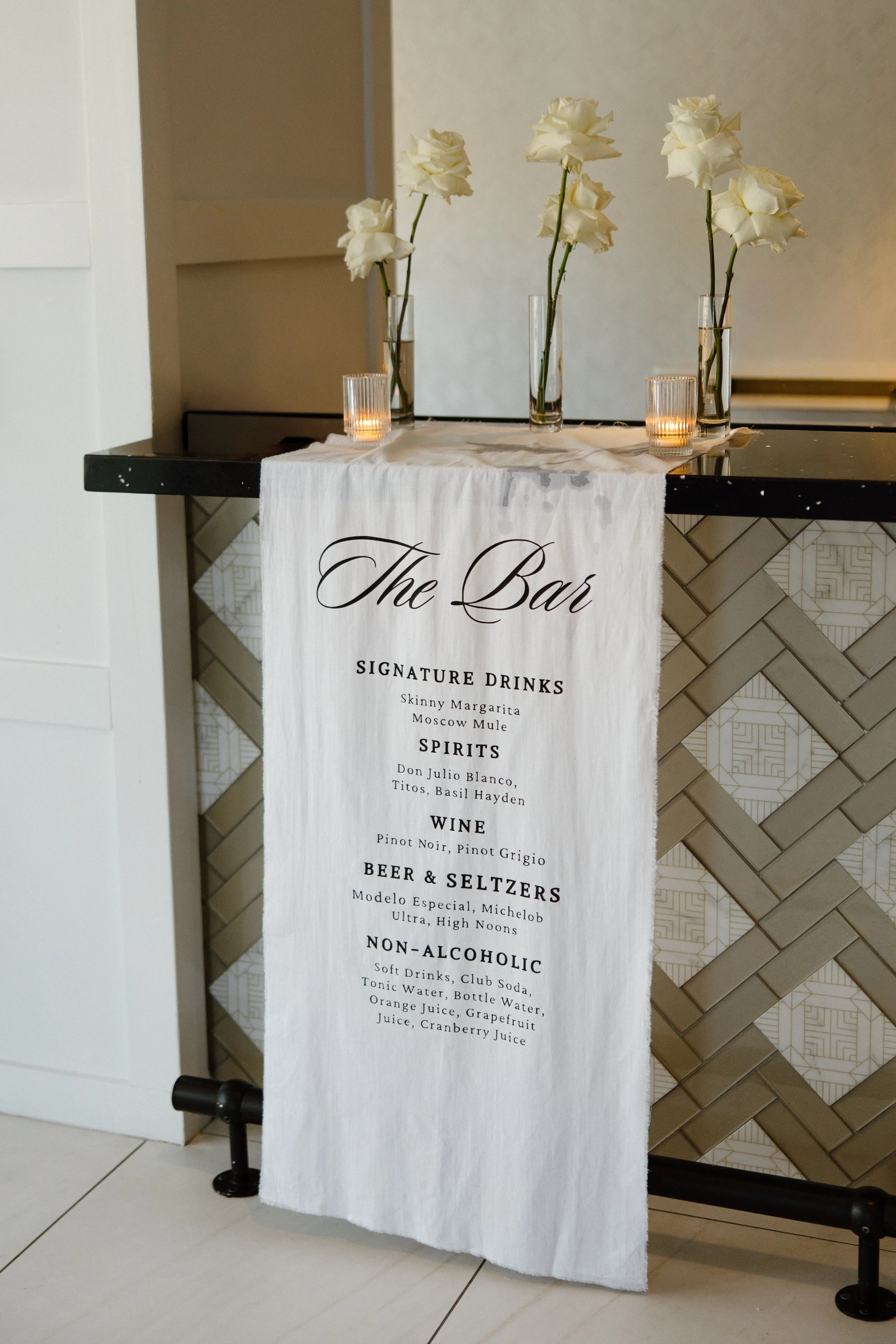 A white cloth sign hanging on a black stand with the words 'The Bar' printed at the top and a list of available drinks below. The drinks include cocktails, spirits, wines, beers, and non-alcoholic beverages, along with their various options.