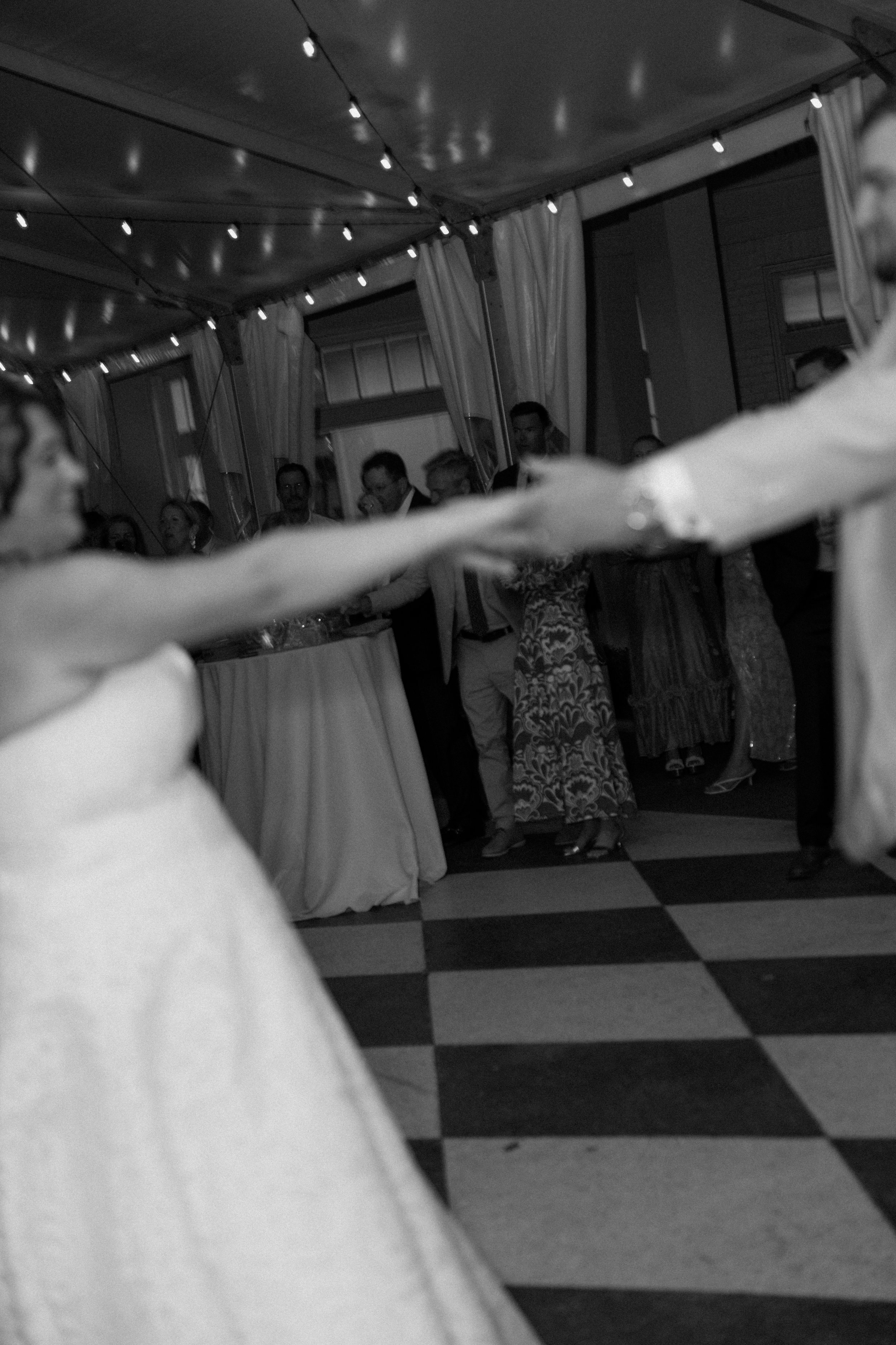 People dancing at a celebration or party, with string lights hanging from the ceiling and curtains on the windows.