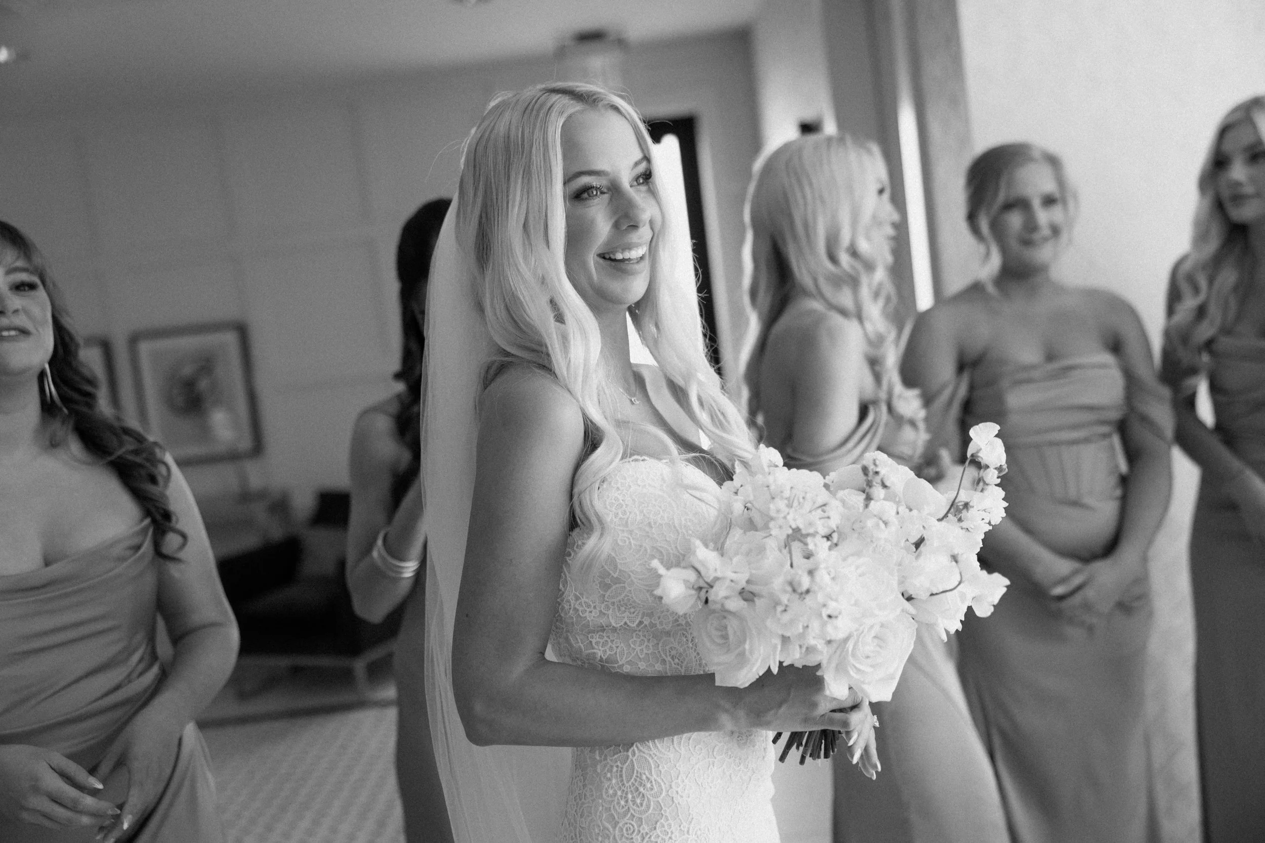 A bride dressed in a lace wedding gown holding a bouquet of white flowers, smiling as she stands with her bridesmaids in a room.