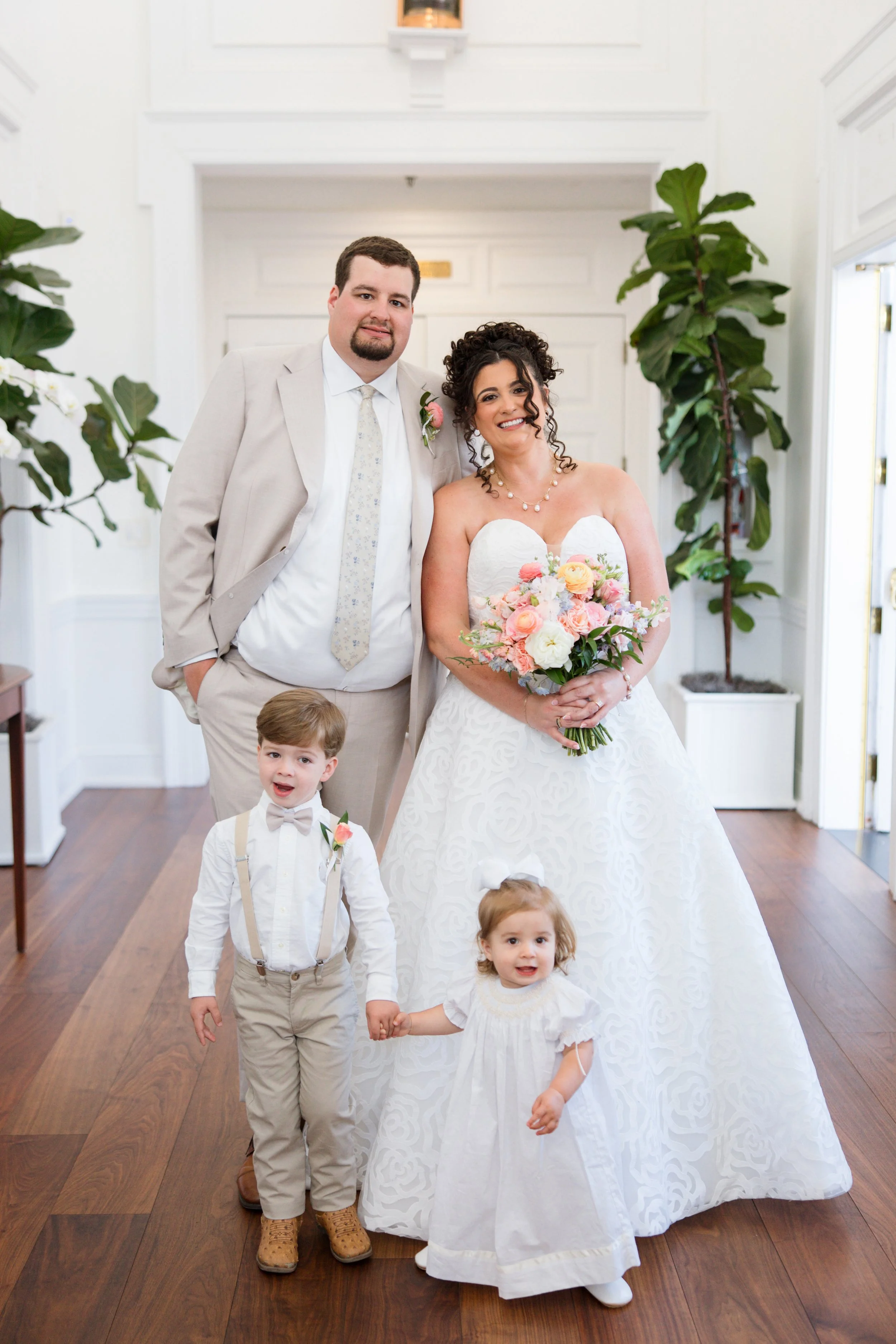 A group of six people, including a bride in a white wedding dress holding a bouquet, a groom in a beige suit, and four young children, standing indoors with white walls, large windows, and wooden floors.