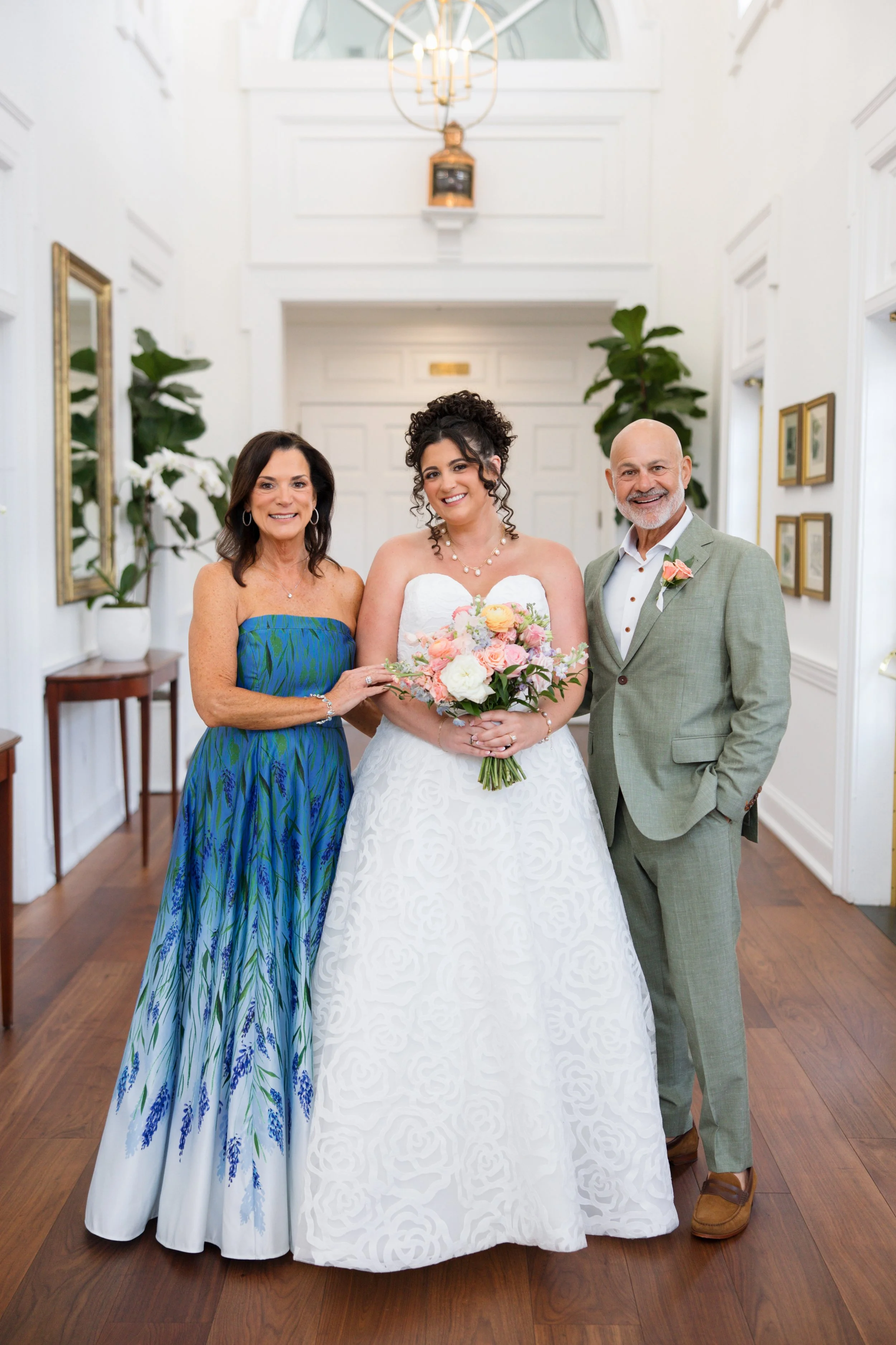 Three people standing together inside a bright, white room with wooden floors, holding a bouquet of flowers, smiling at the camera. The woman in the center is wearing a white wedding dress, flanked by an older woman in a strapless blue dress on her l