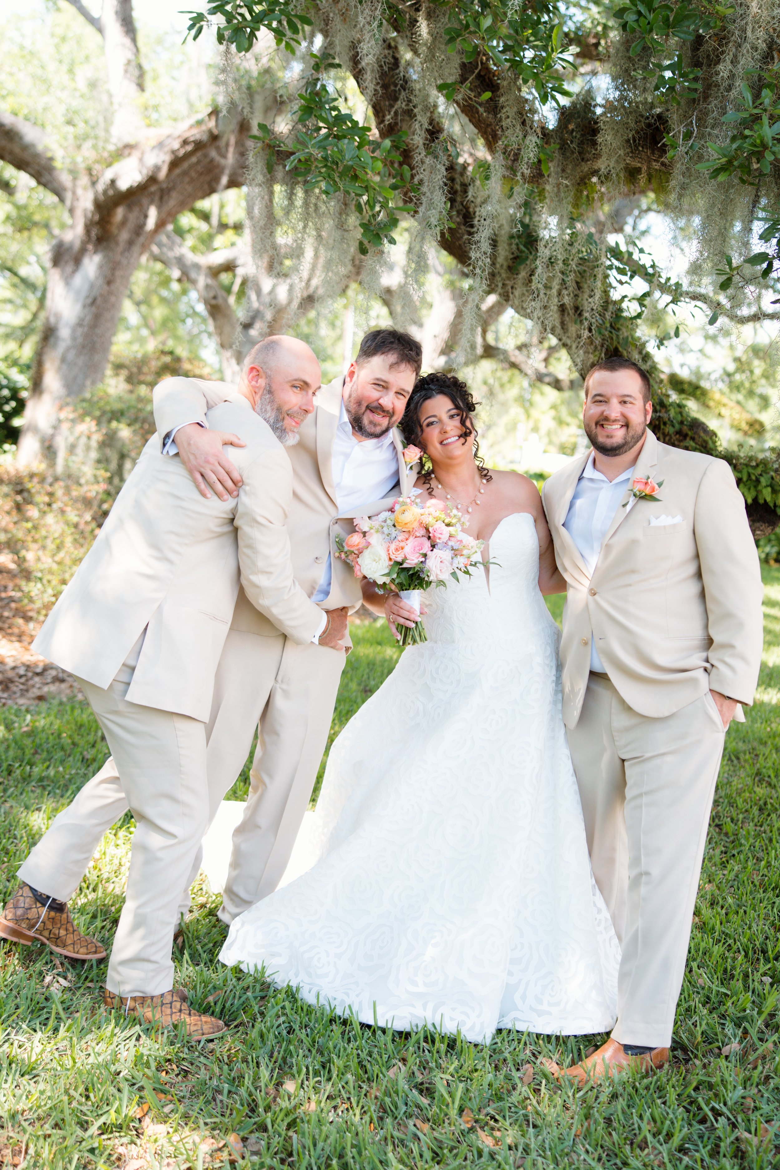 A group of four people dressed in wedding attire outdoors under a tree with hanging moss, smiling and hugging each other, with greenery and trees in the background.