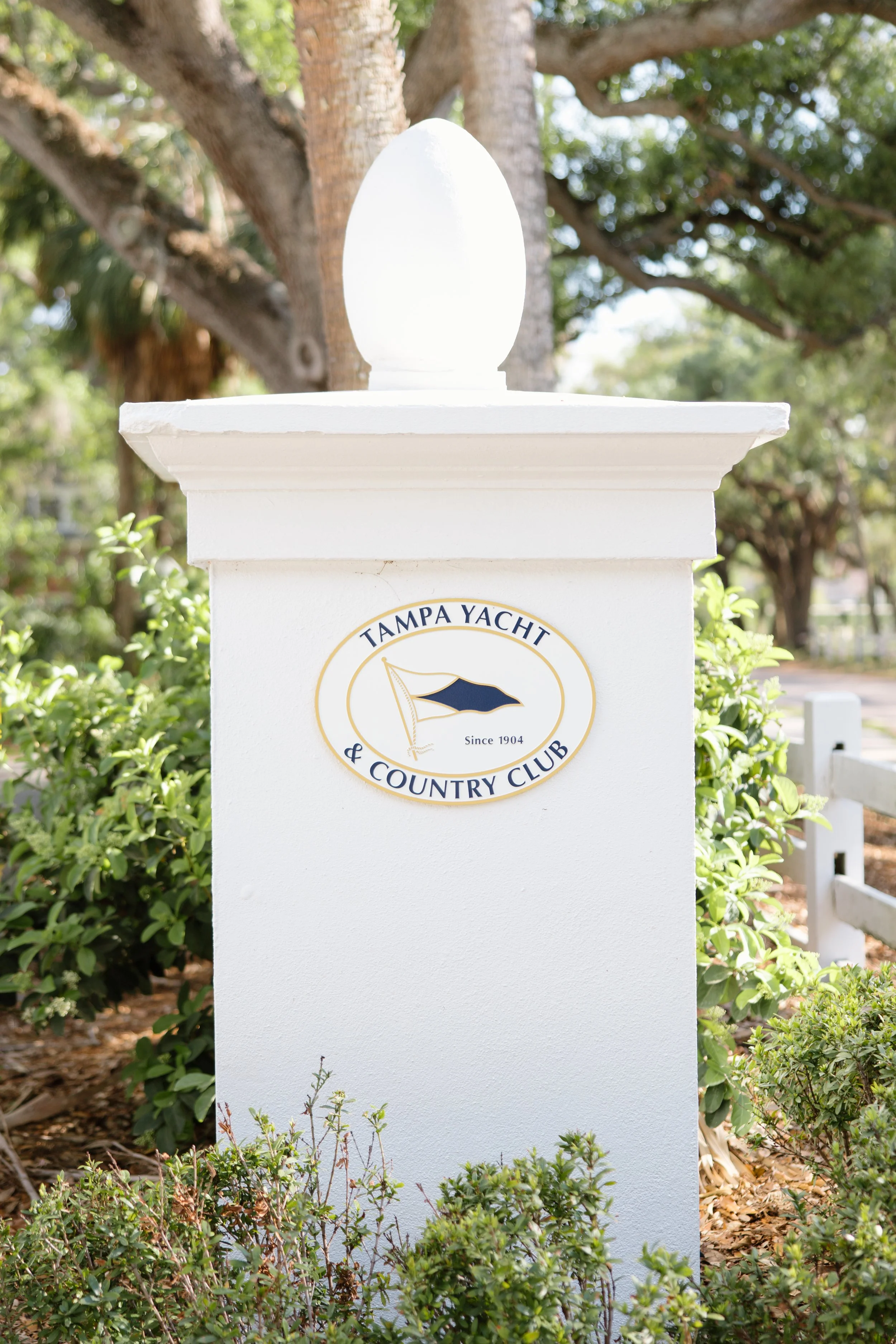 White signpost with a round emblem reading 'Tampa Yacht & Country Club' and a sailboat logo, surrounded by green bushes and trees.