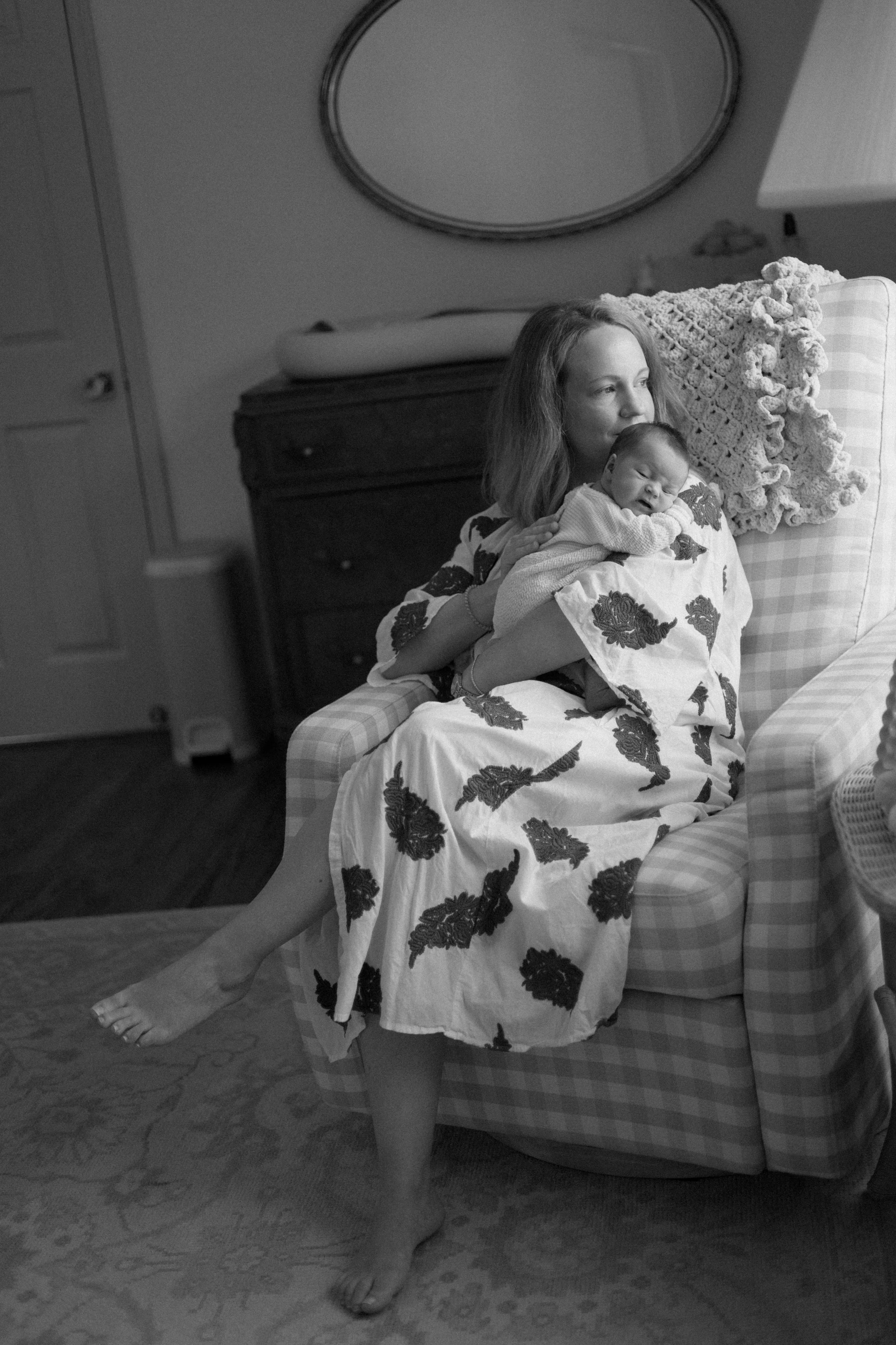 A woman sitting on an armchair holding a baby. The woman is dressed in a floral dress, and the baby is wrapped in a blanket. There is a mirror on the wall behind them and a side table with a bowl on it. The woman is barefoot, with one foot on the ground and the other resting on the edge of the armchair.