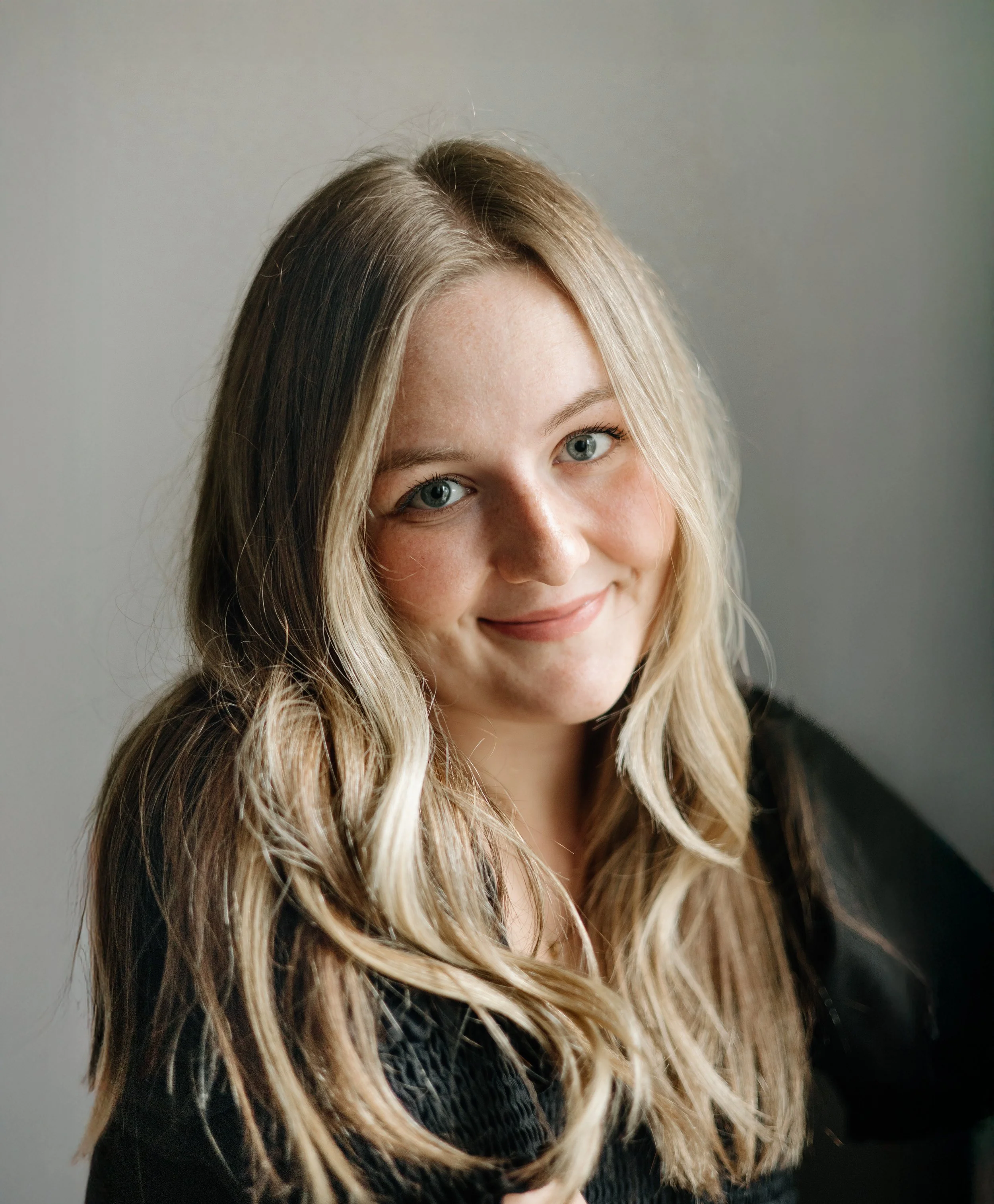A woman with long wavy blonde hair and bright blue eyes smiling slightly, wearing a black top, in front of a plain light gray background.