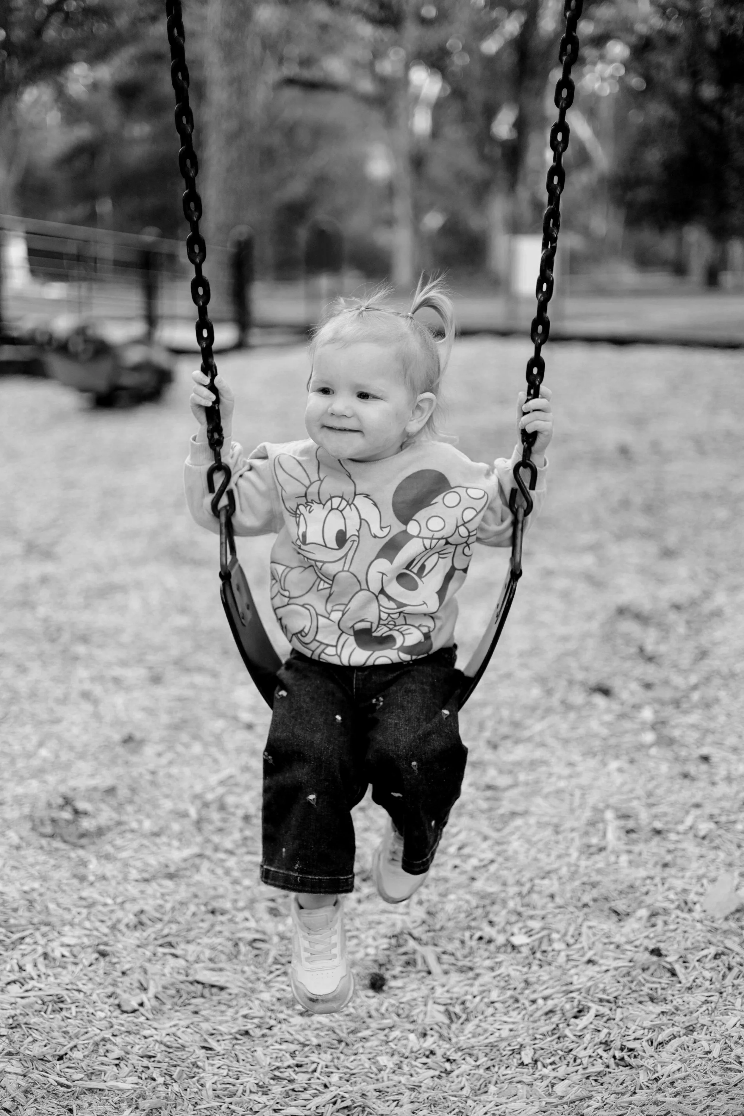 A young child, wearing a Mickey Mouse sweatshirt, black pants, and sneakers, is smiling and running on a playground swing with chains, surrounded by wood chips.