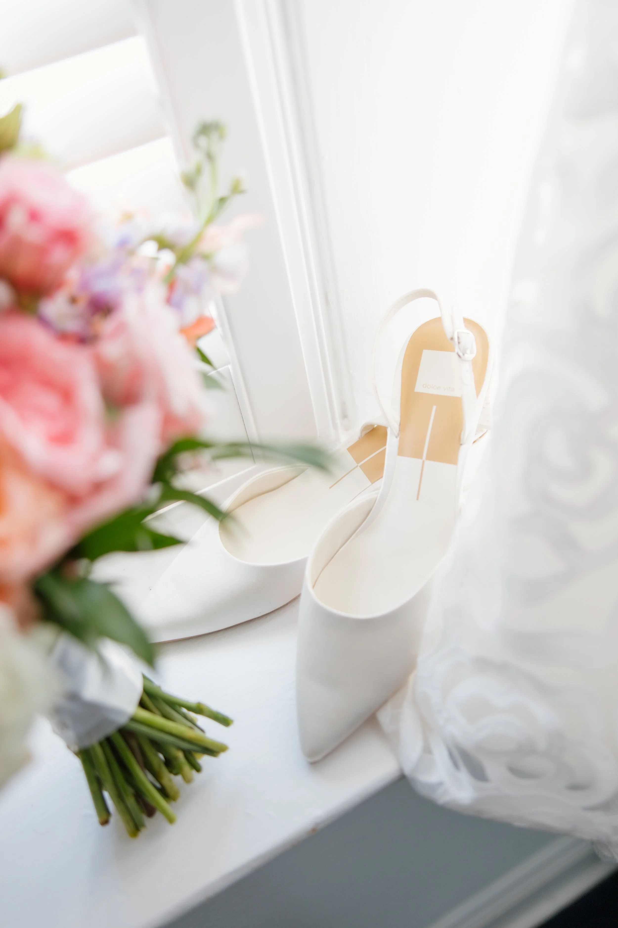 A bouquet of pink, white, and purple flowers resting on a white surface with white high heels beside it. In the background, a white door with a beige tag hanging from the doorknob.