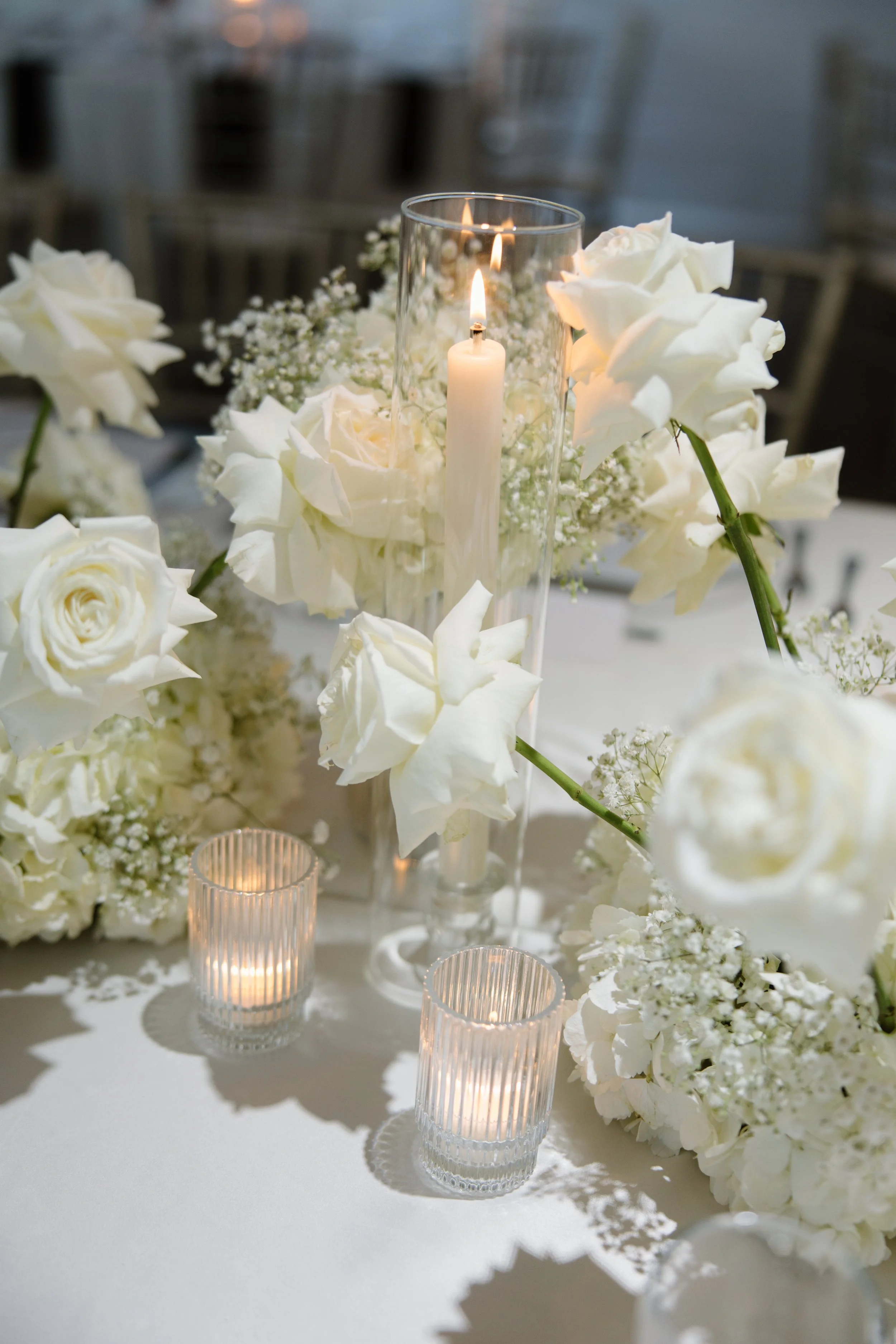 A centerpiece of white roses, baby's breath, and hydrangeas with candles in clear glass holders on a white table at an event.