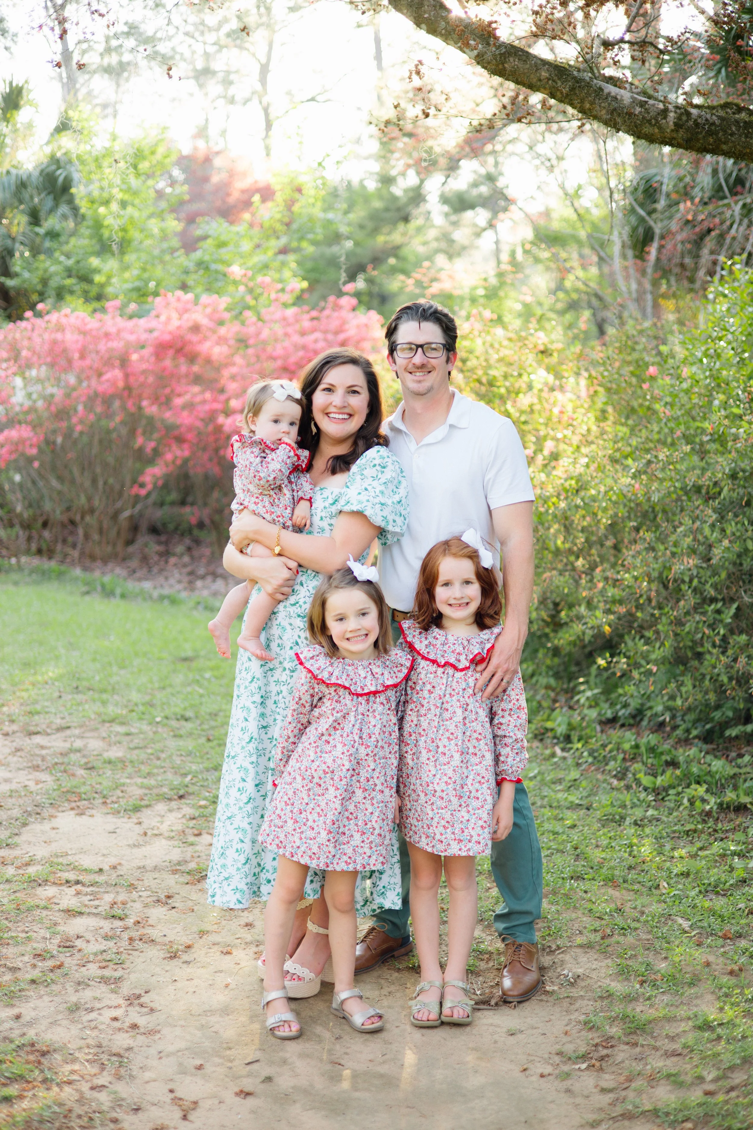 A family of five posing outdoors in a park during daytime, surrounded by colorful trees and pink flowering bushes, all smiling and dressed in casual summer clothes.