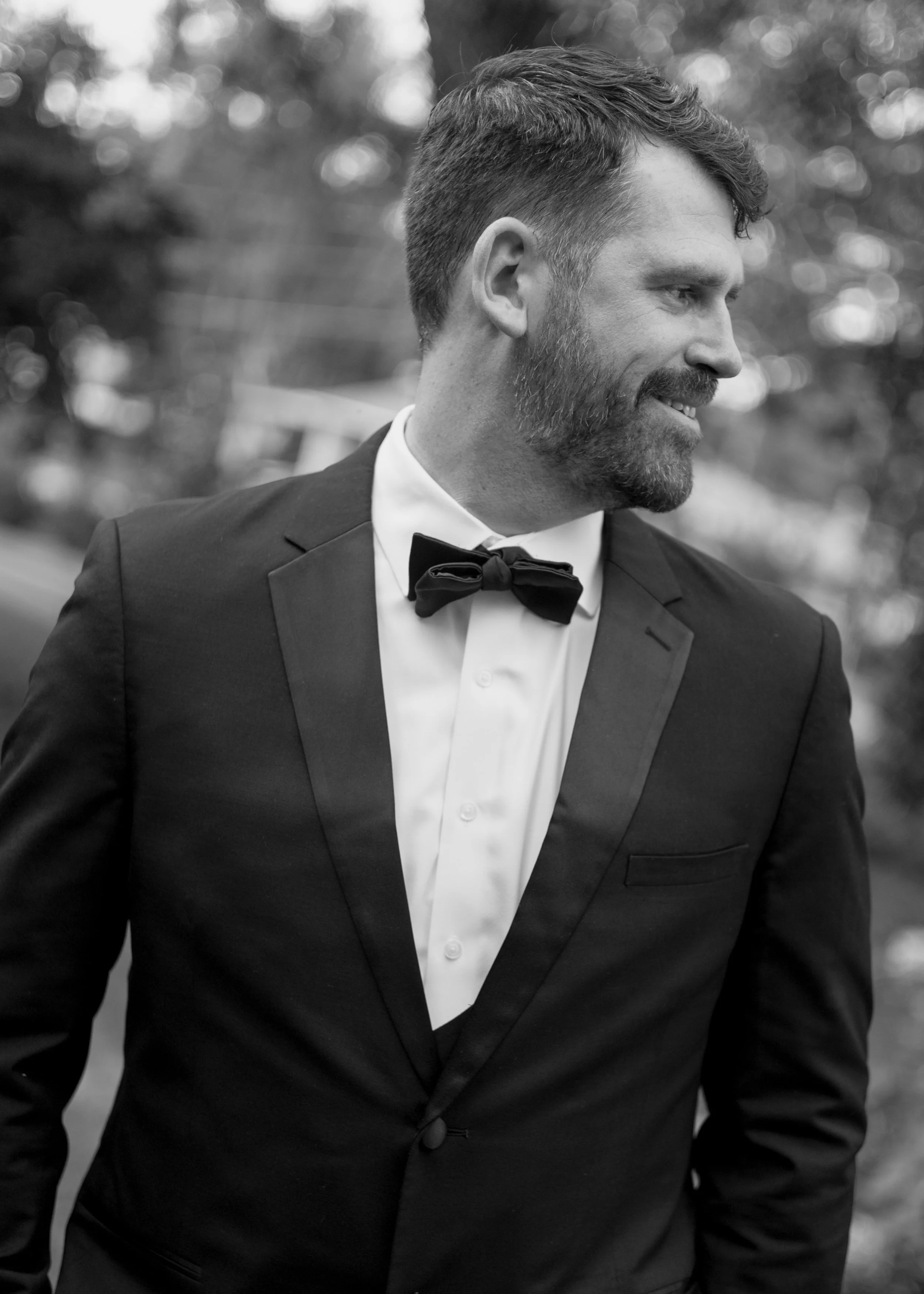 A man dressed in a tuxedo with a bow tie, smiling and looking to the right, outdoors with blurred trees in the background.