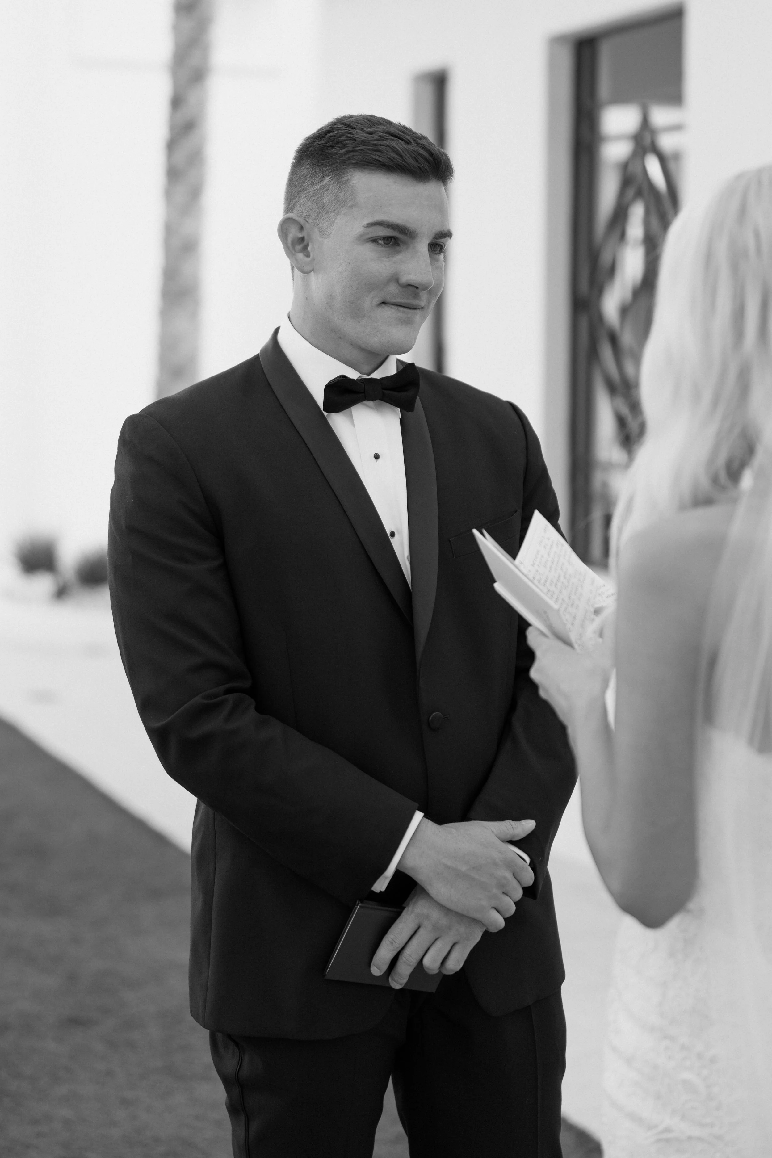 A young man in a tuxedo with a bow tie holding a notebook, engaged in a conversation with a woman whose back is to the camera.